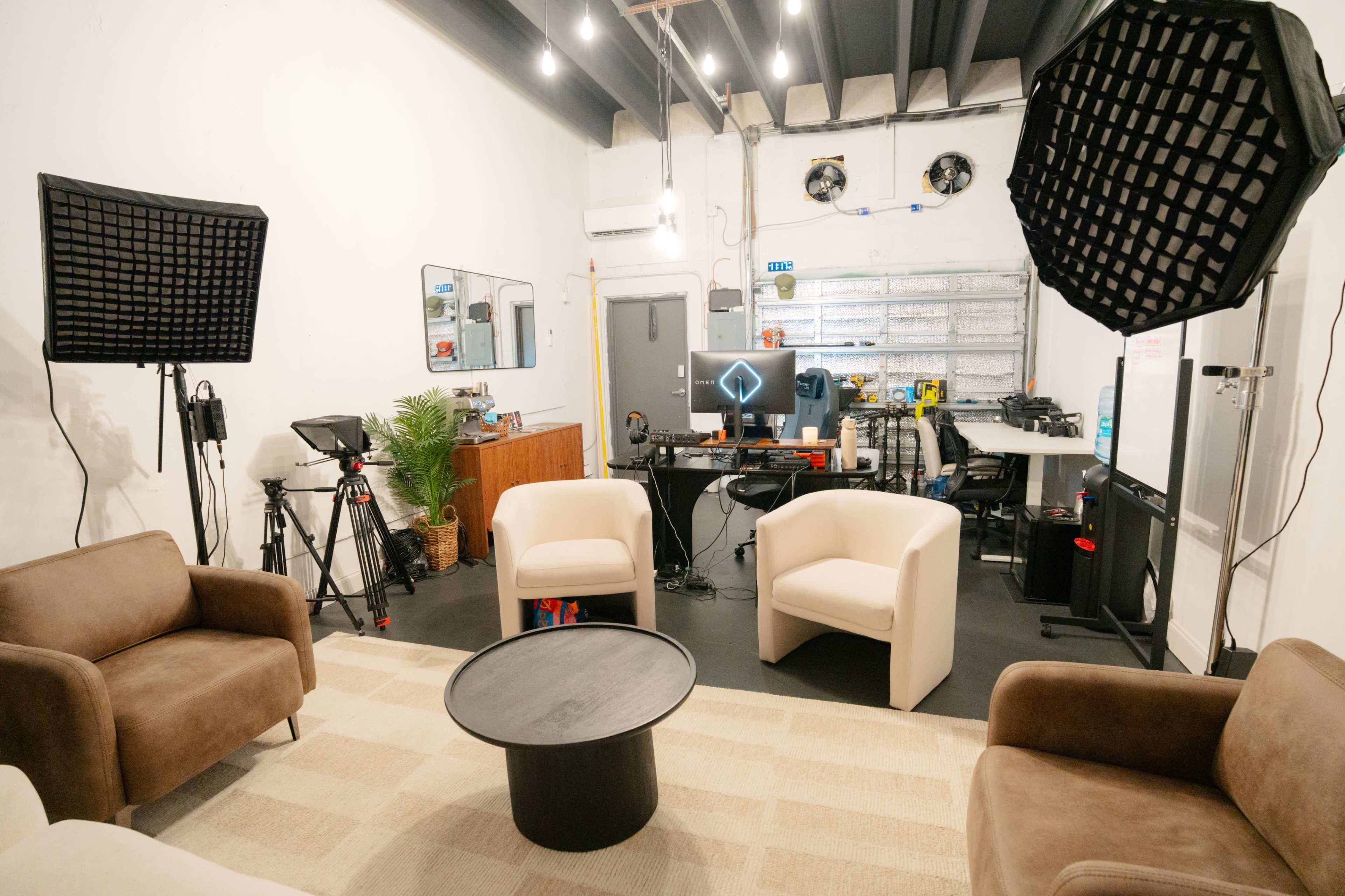 The image shows a modern studio space with brown and cream armchairs arranged around a black circular table, cameras on tripods, and lighting equipment set up in a well-lit environment.