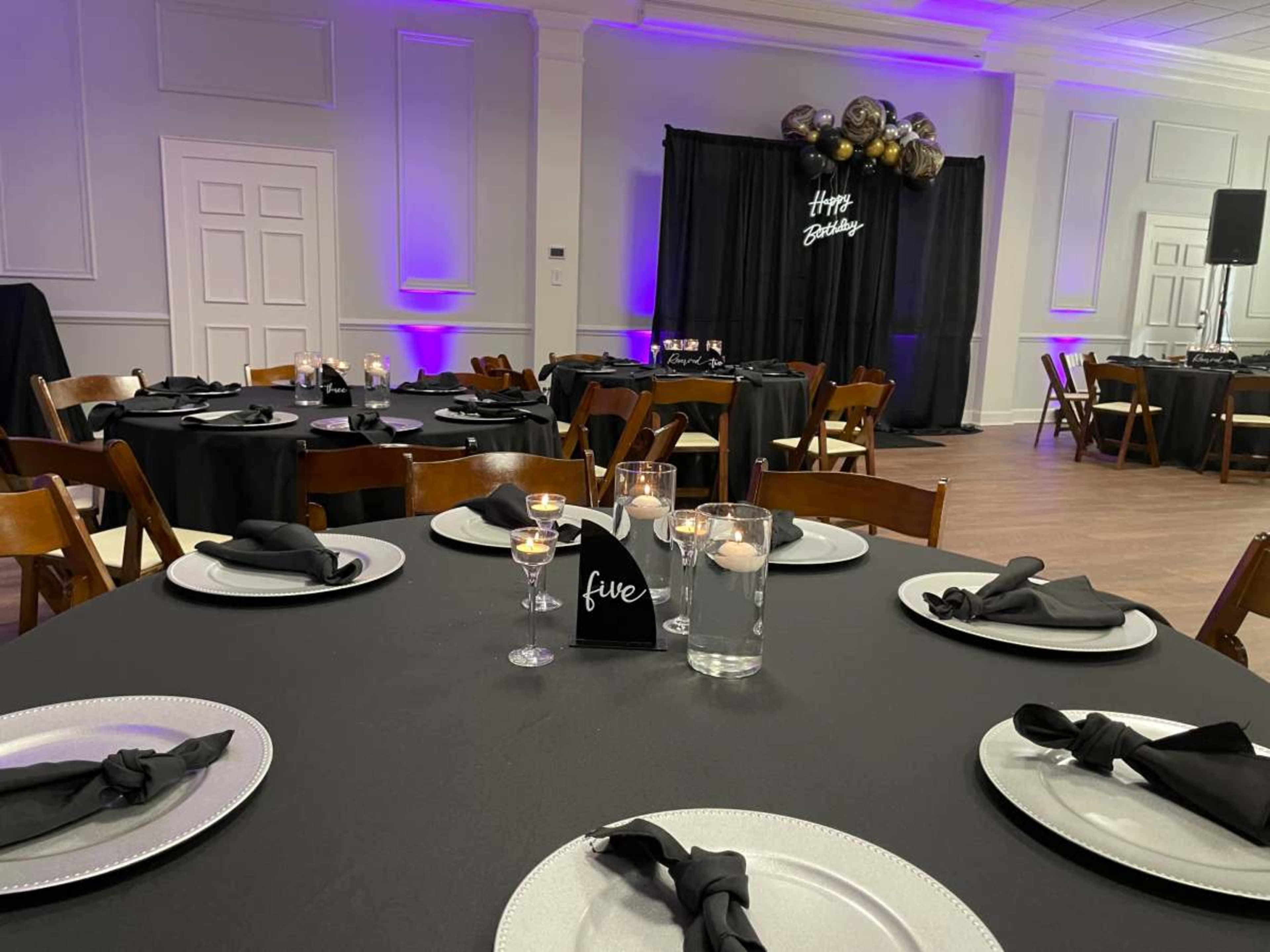 A decorated event space with black tablecloths, neatly set tables featuring silver chargers and napkins, and a backdrop with "Happy Birthday" in a festive arrangement.