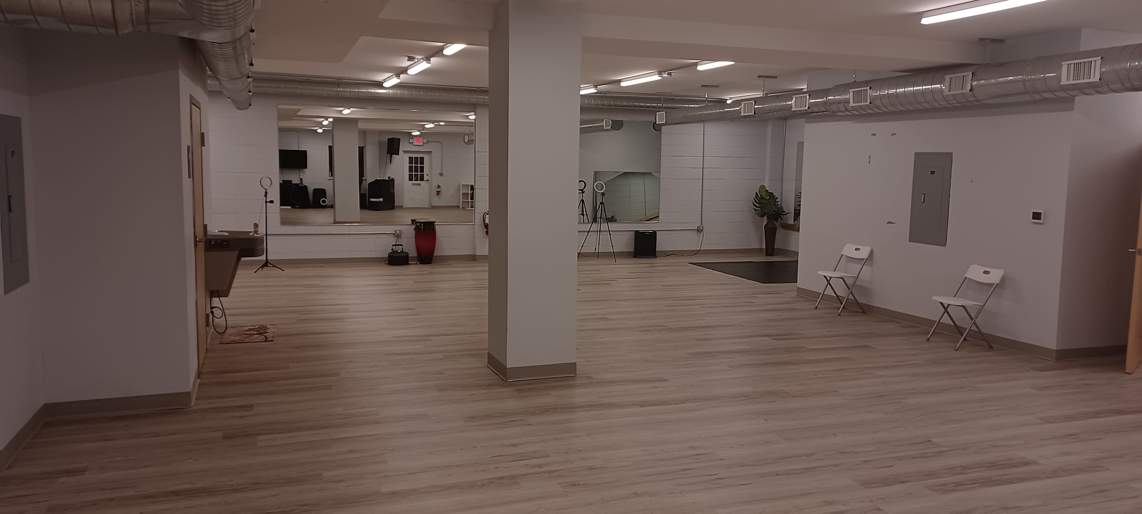 The image shows a spacious, empty room with light-colored wooden flooring and basic furnishings, including chairs and wall-mounted mirrors.