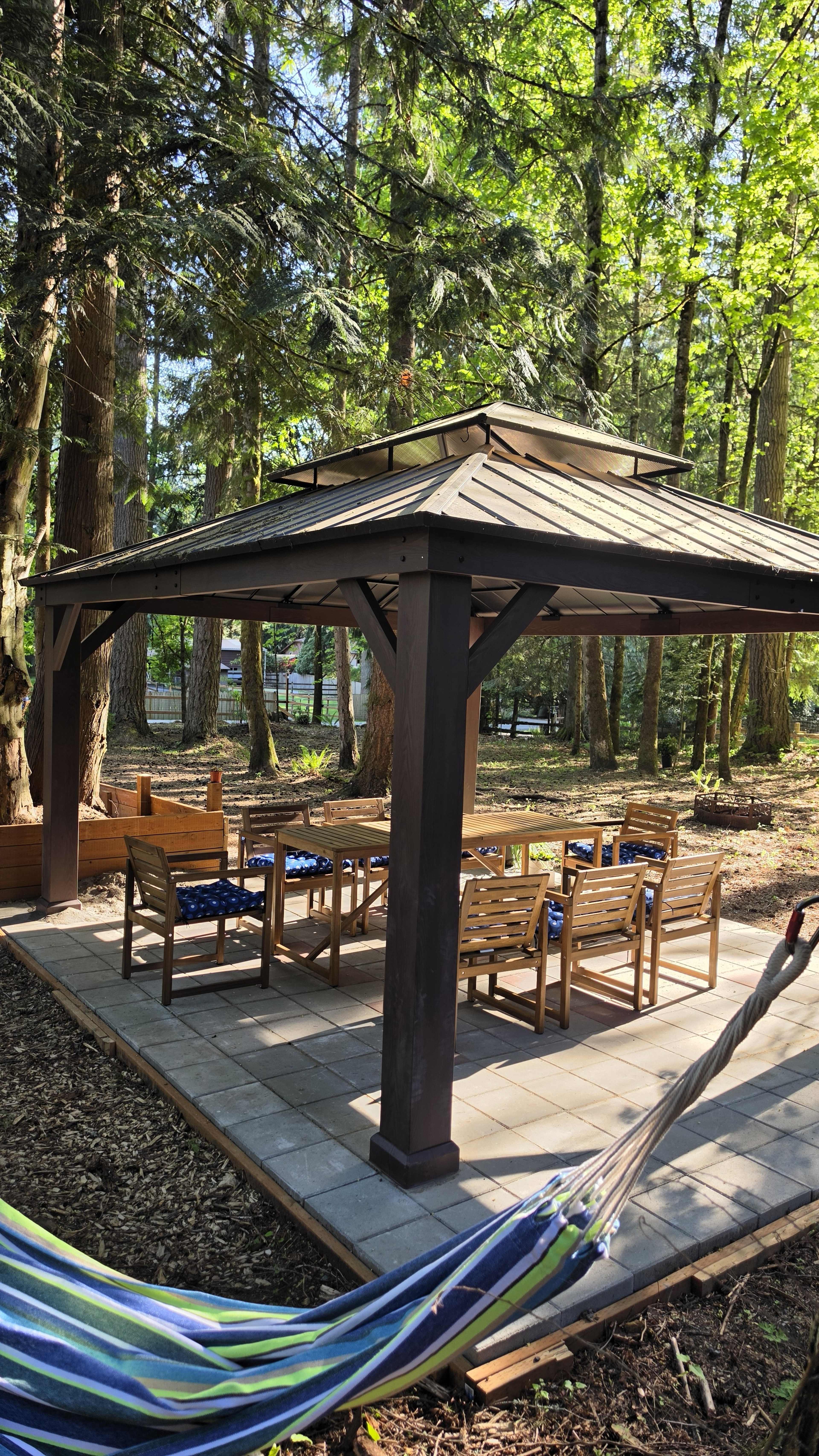 A wooden gazebo with a tiled floor contains several wooden chairs and tables, surrounded by trees in a wooded area.