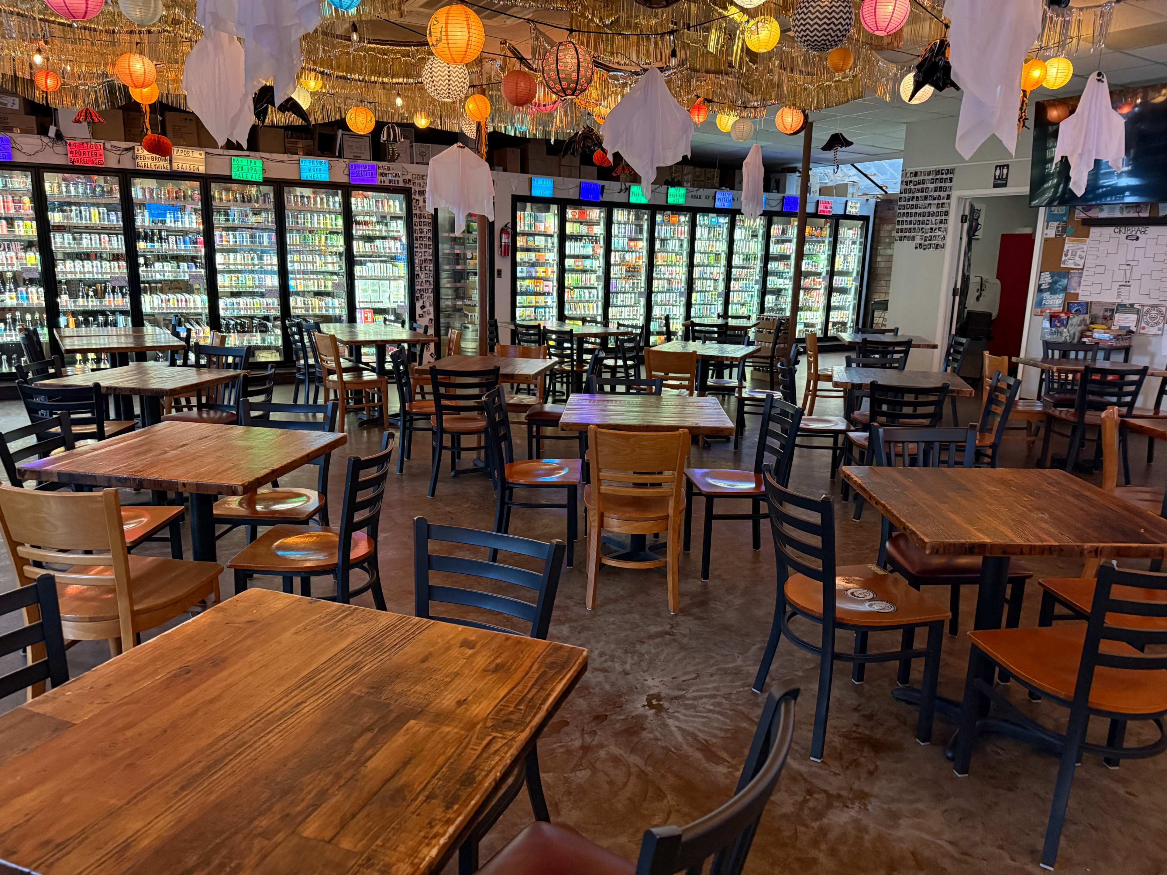 The image shows a spacious indoor dining area with wooden tables and chairs, decorated with colorful hanging lights and filled with beverage coolers along the walls.