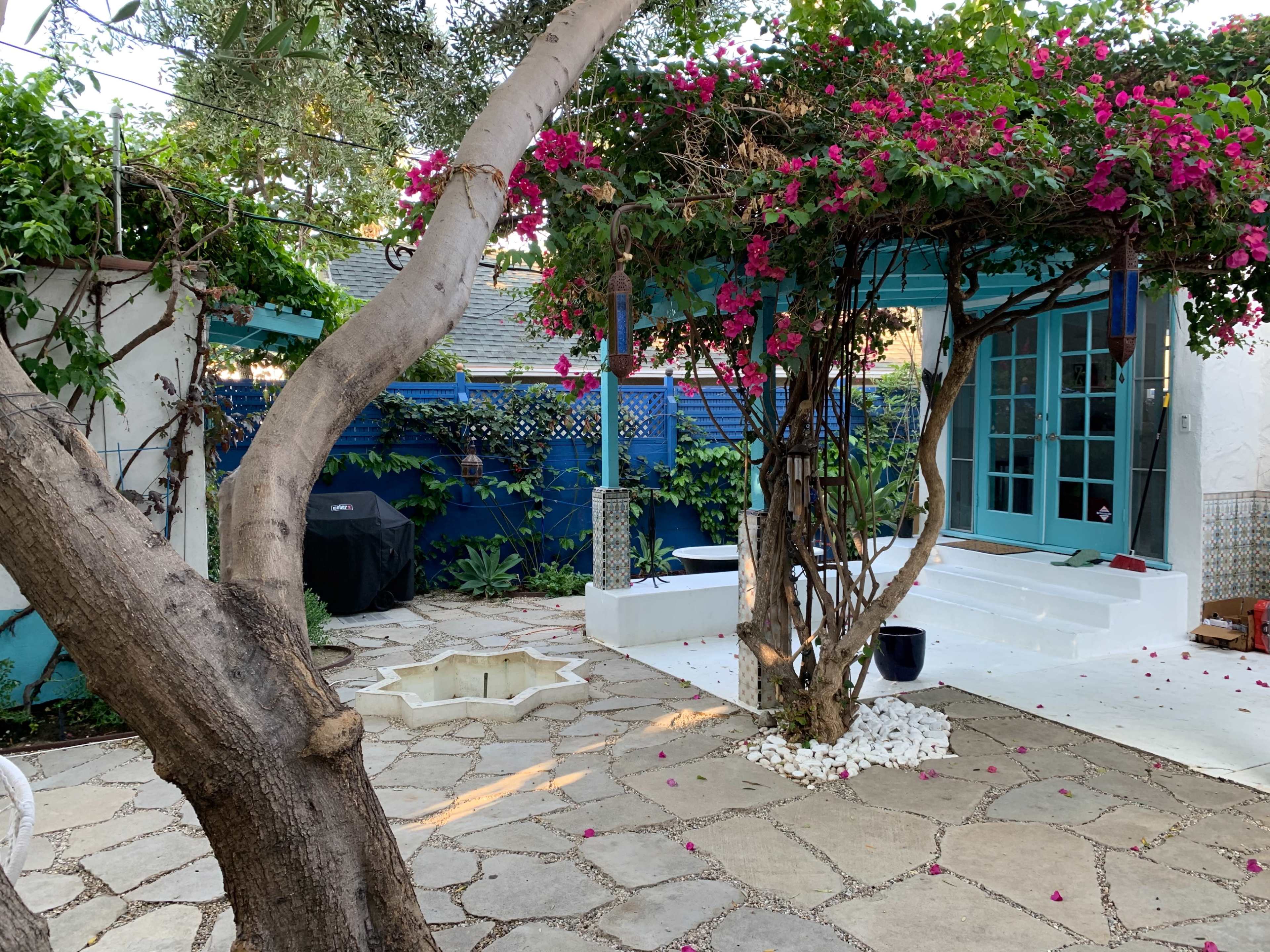 The scene shows a landscaped outdoor space with a stone patio, a blue structure, blooming bougainvillea, and a circular stone basin surrounded by white stones.