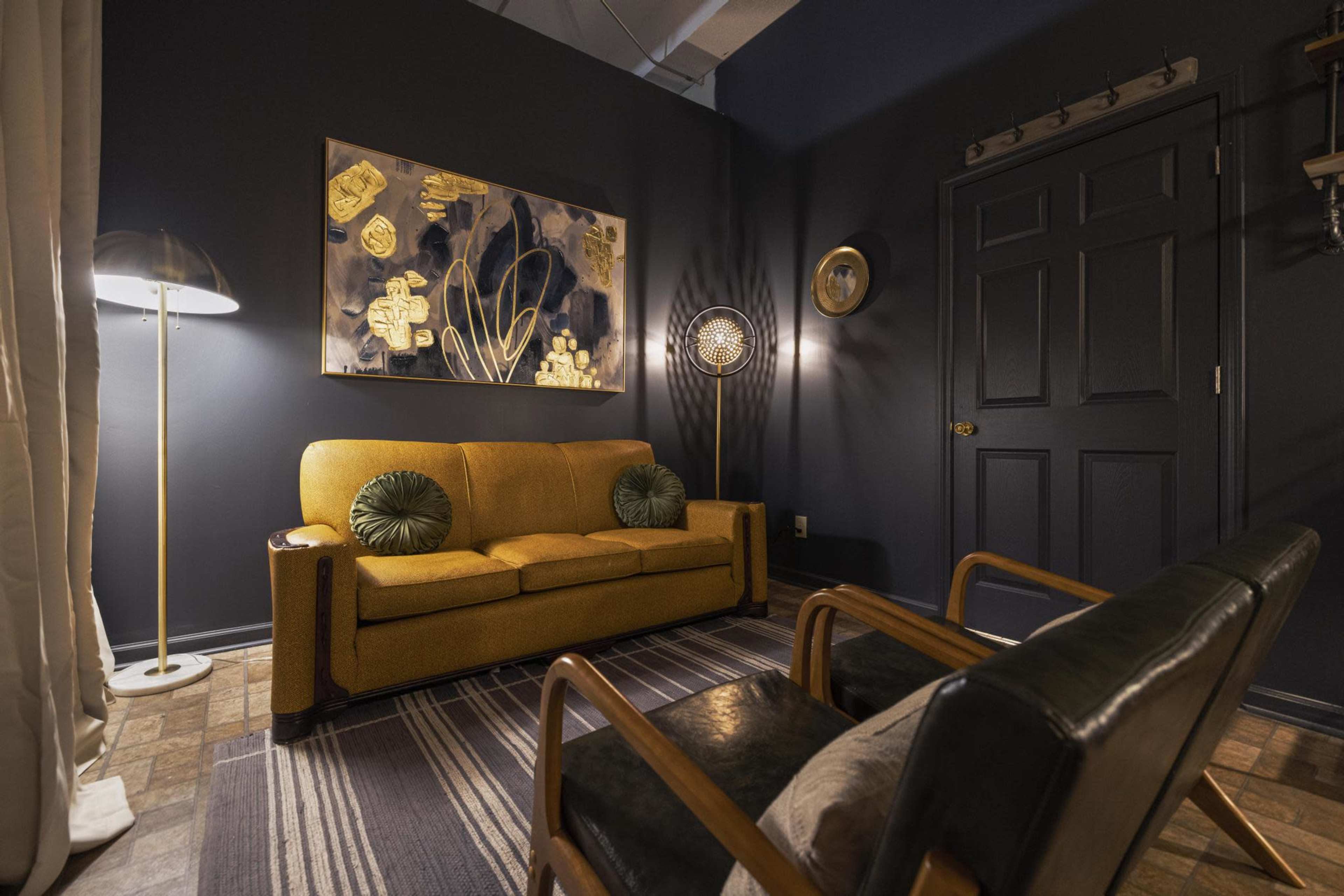A cozy living room corner with a yellow sofa, two black armchairs, and a dark blue wall featuring a decorative painting.