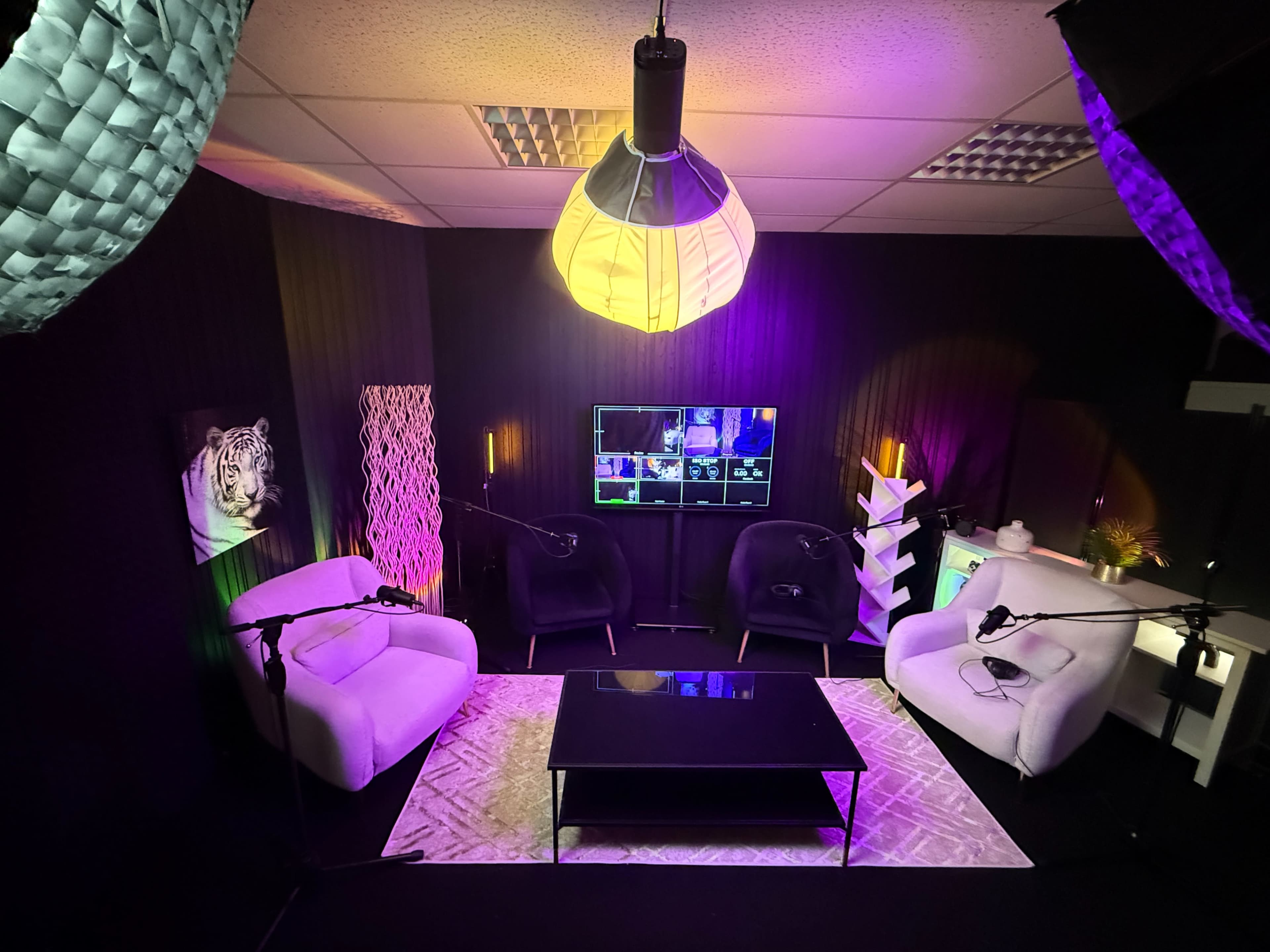 The image shows a modern studio setup featuring two white chairs, a black coffee table, and a large screen displaying multiple camera angles, all surrounded by colorful lighting and decorative elements.