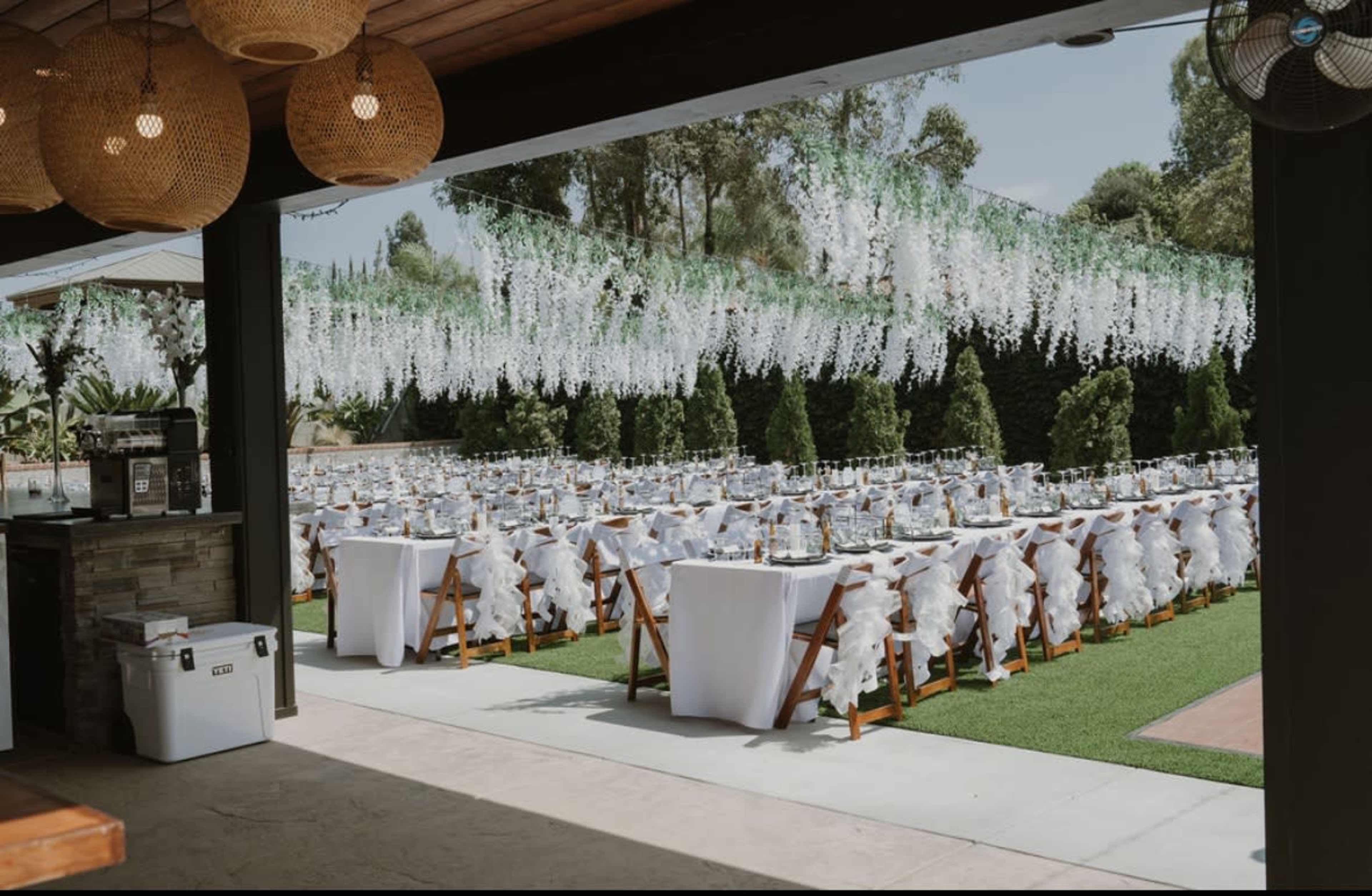 An outdoor event space is set up with numerous tables draped in white tablecloths and decorated with elegant floral arrangements under hanging greenery.