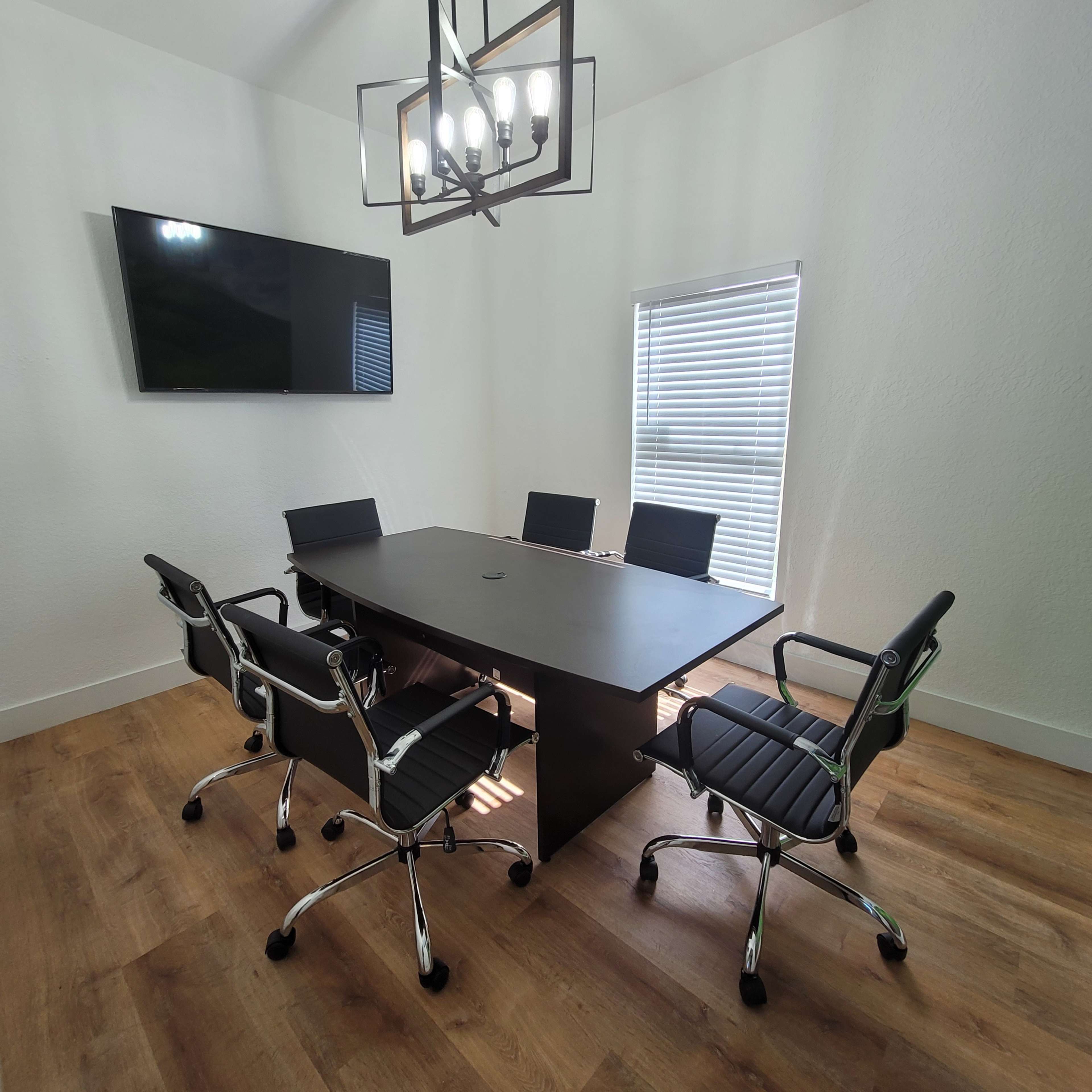 A modern conference room features a large rectangular table surrounded by six black ergonomic chairs, with a wall-mounted television and a window providing natural light.