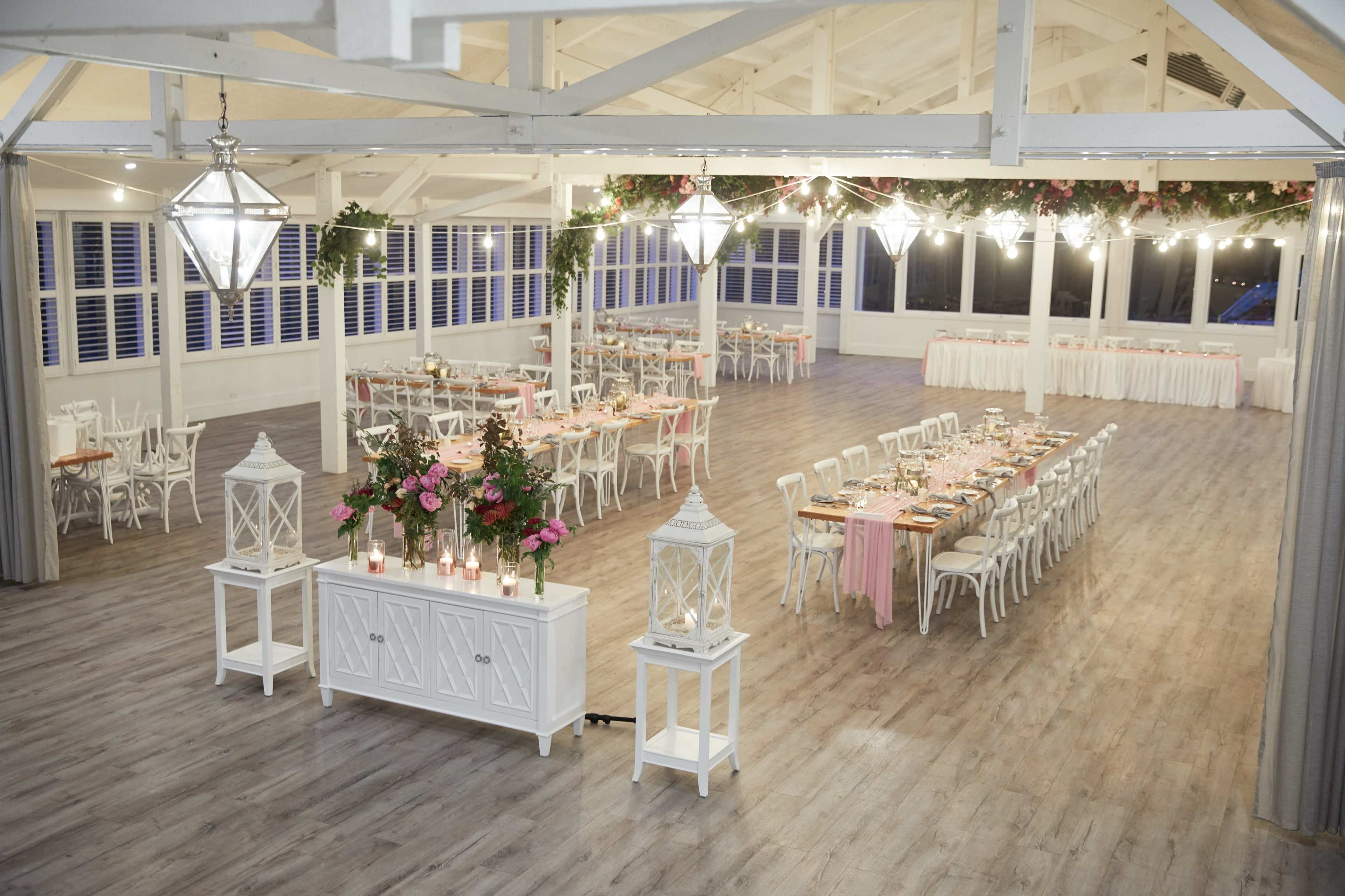 The image shows a spacious event hall styled for a banquet, featuring long tables decorated with floral arrangements and soft lighting.