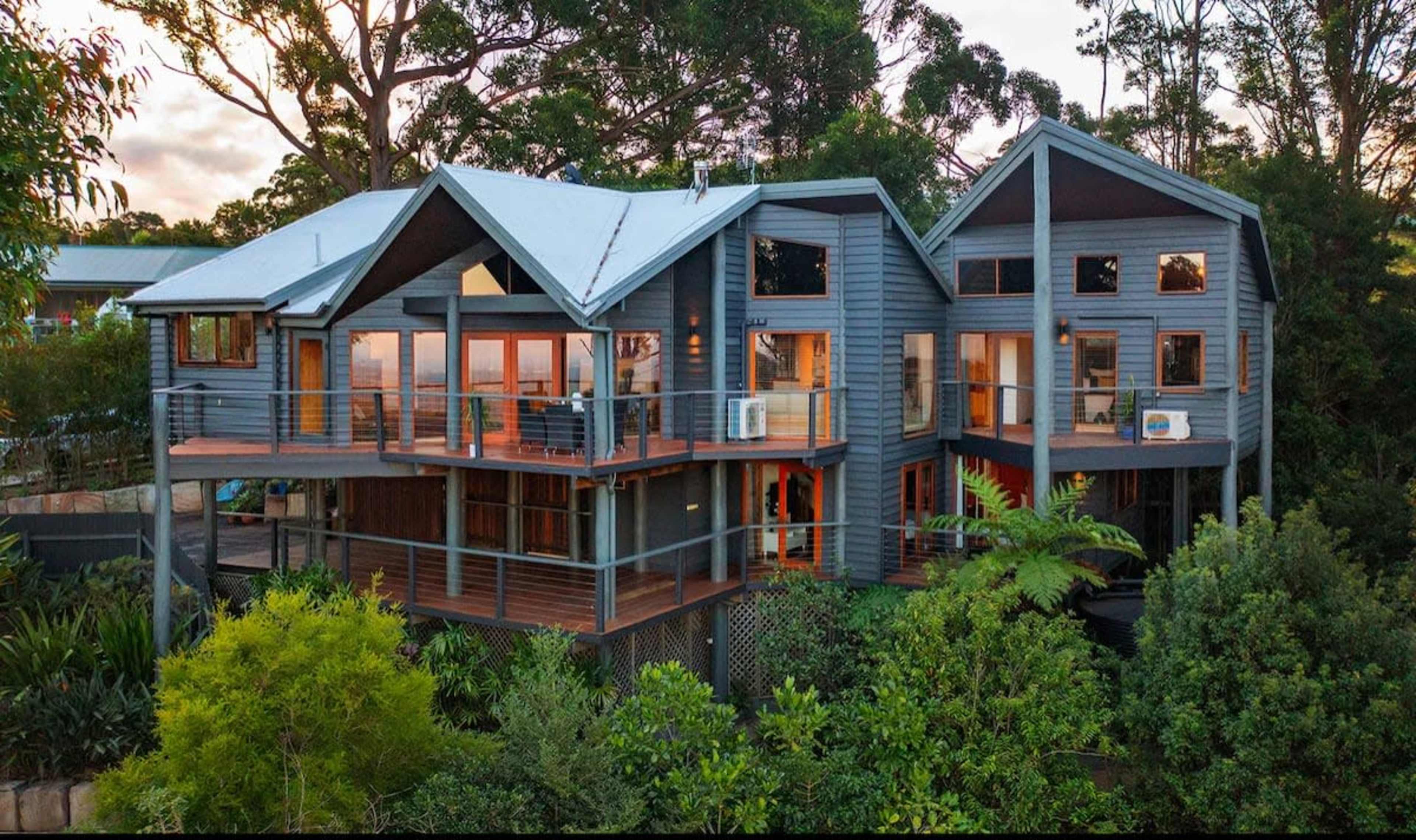 The image shows a modern two-story wooden house surrounded by greenery, featuring large windows and a multi-gabled roof.