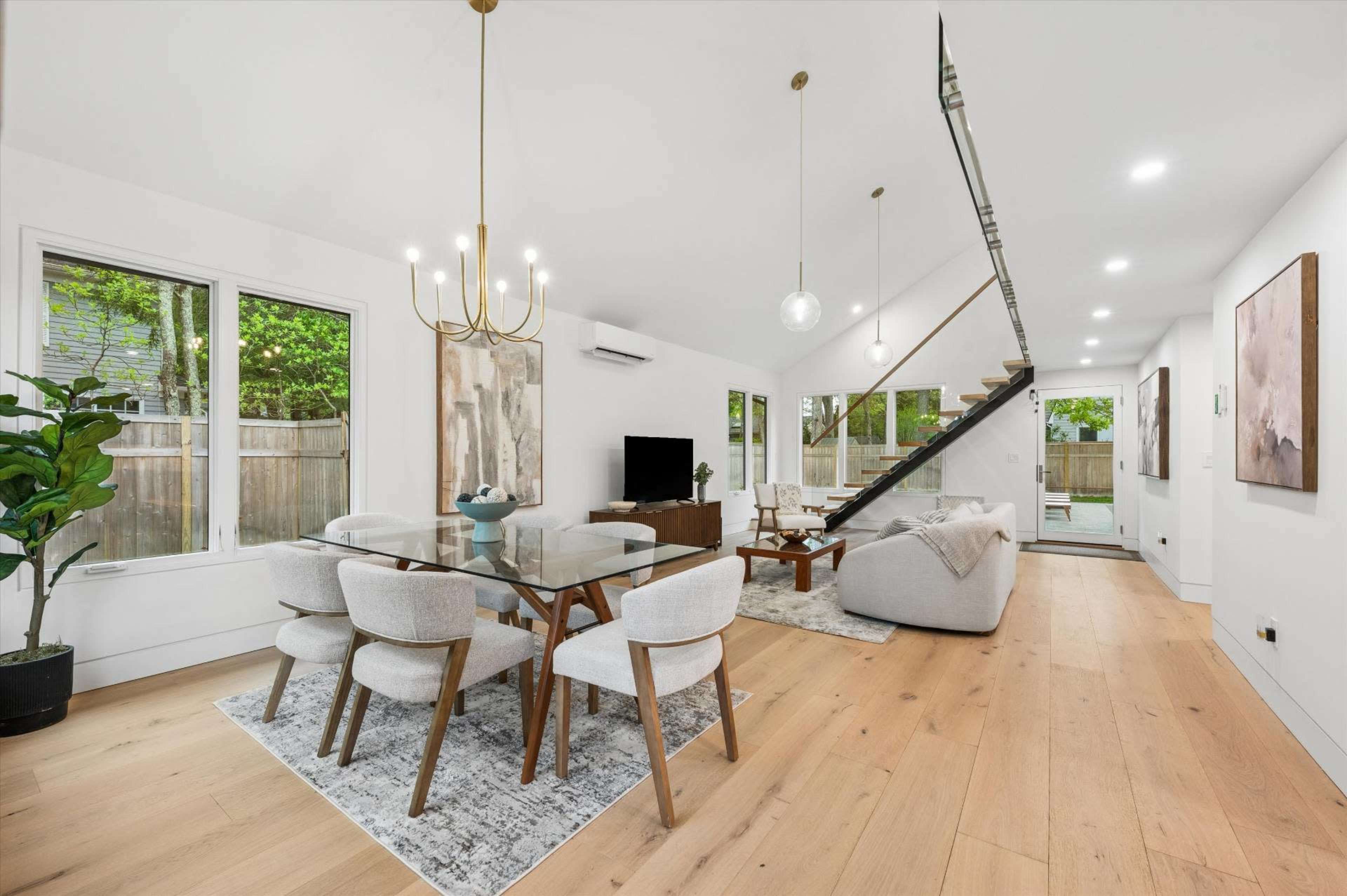 The image shows a modern, open-plan living and dining area with a glass dining table, light-colored furniture, and large windows allowing natural light.