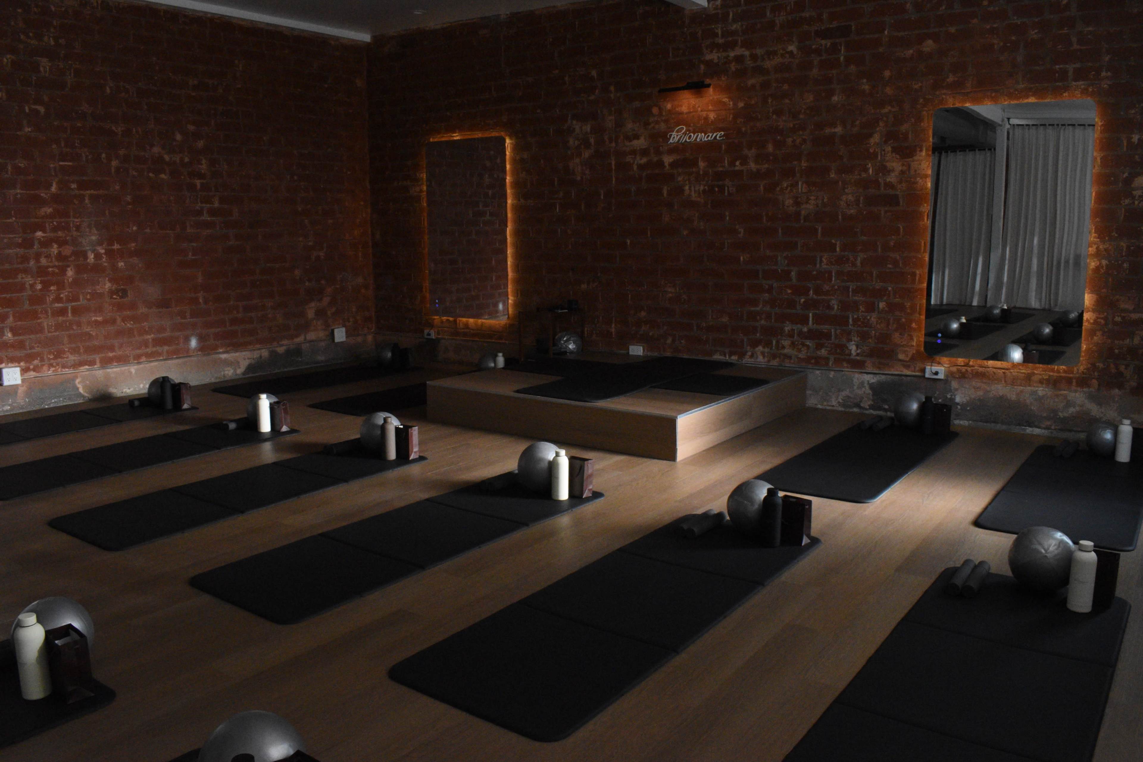 The image shows a dimly lit yoga studio with several mats arranged in rows, a raised platform at the back, and mirrors reflecting the brick walls.