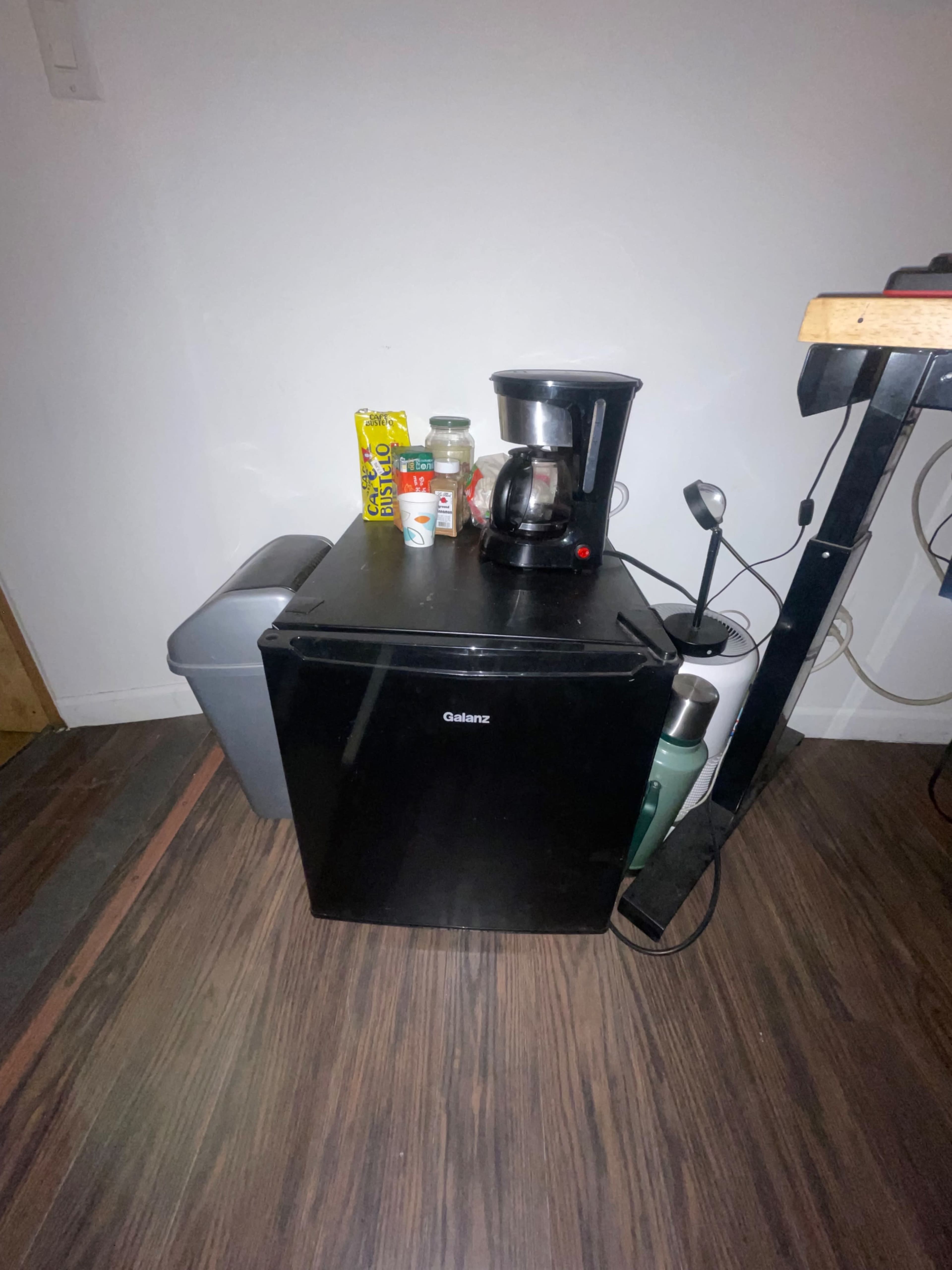 A small black refrigerator sits on a wooden floor next to a trash can and a coffee maker, with some items on top including a box of cereal and a bottle.