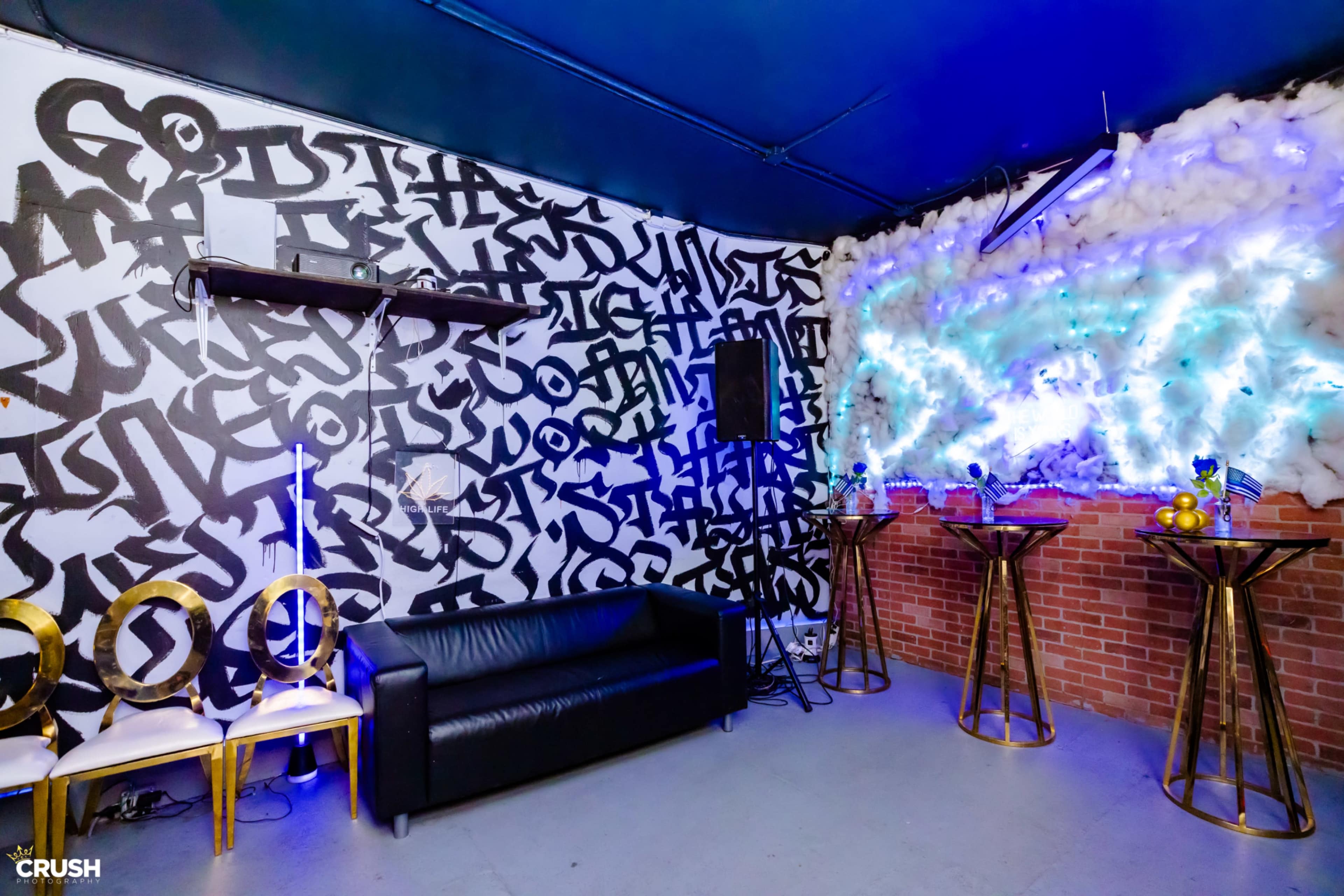 The image shows a modern lounge area with a black-and-white graffiti-style wall, a black sofa, and bar tables decorated with colorful items.