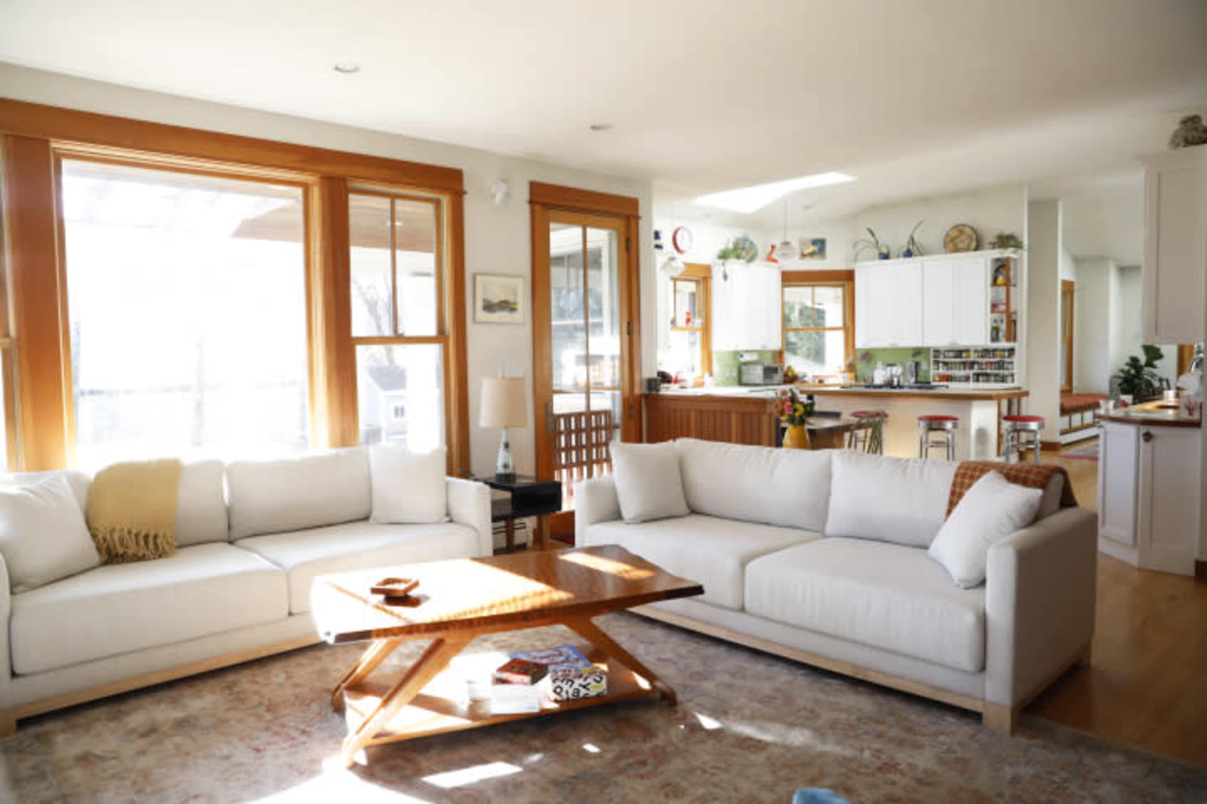 A bright living room features two white sofas arranged around a wooden coffee table on a patterned rug, with large windows illuminating the space.