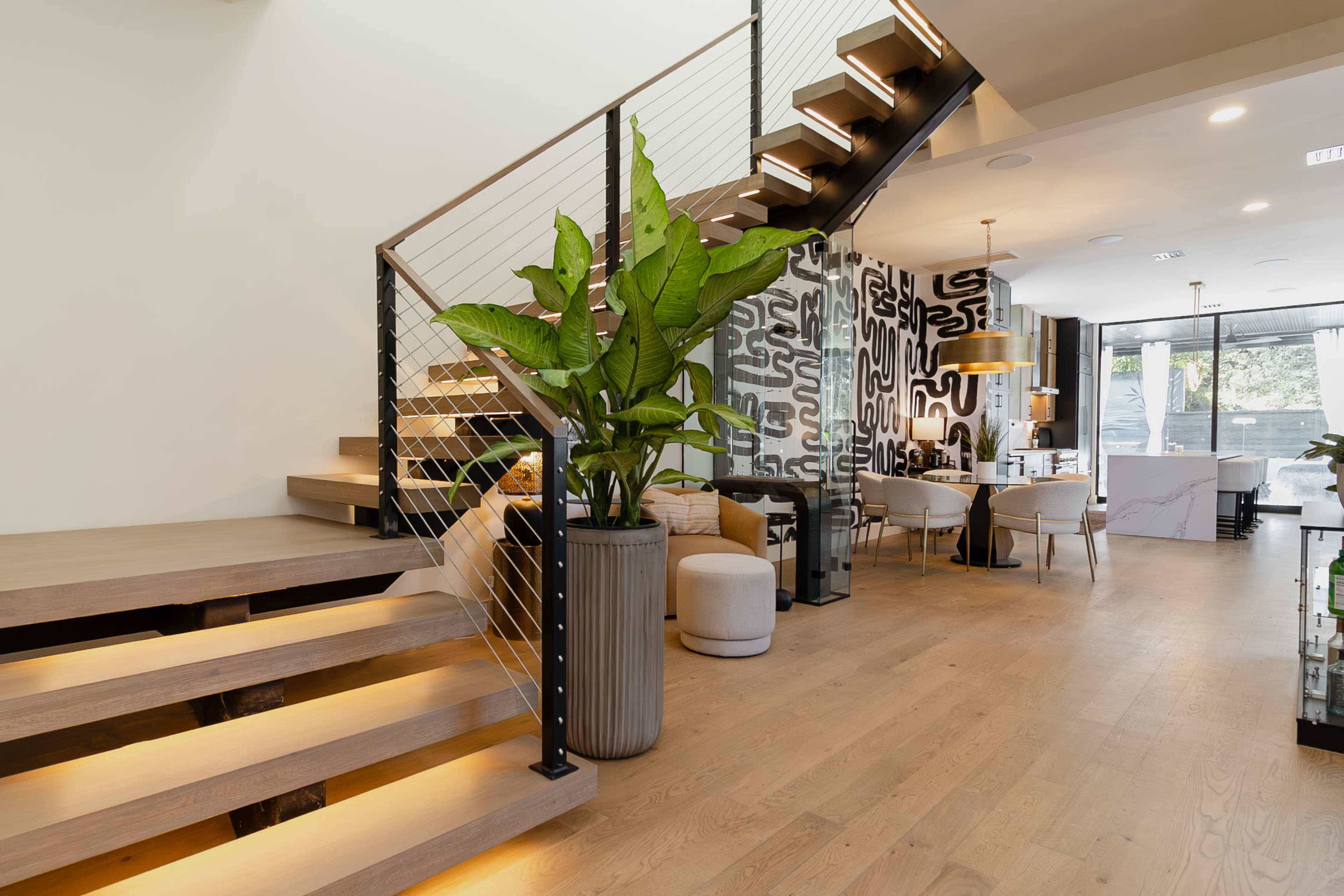 A modern interior showcases a staircase with wooden steps and a black railing, accompanied by a large potted plant and a dining area in the background.
