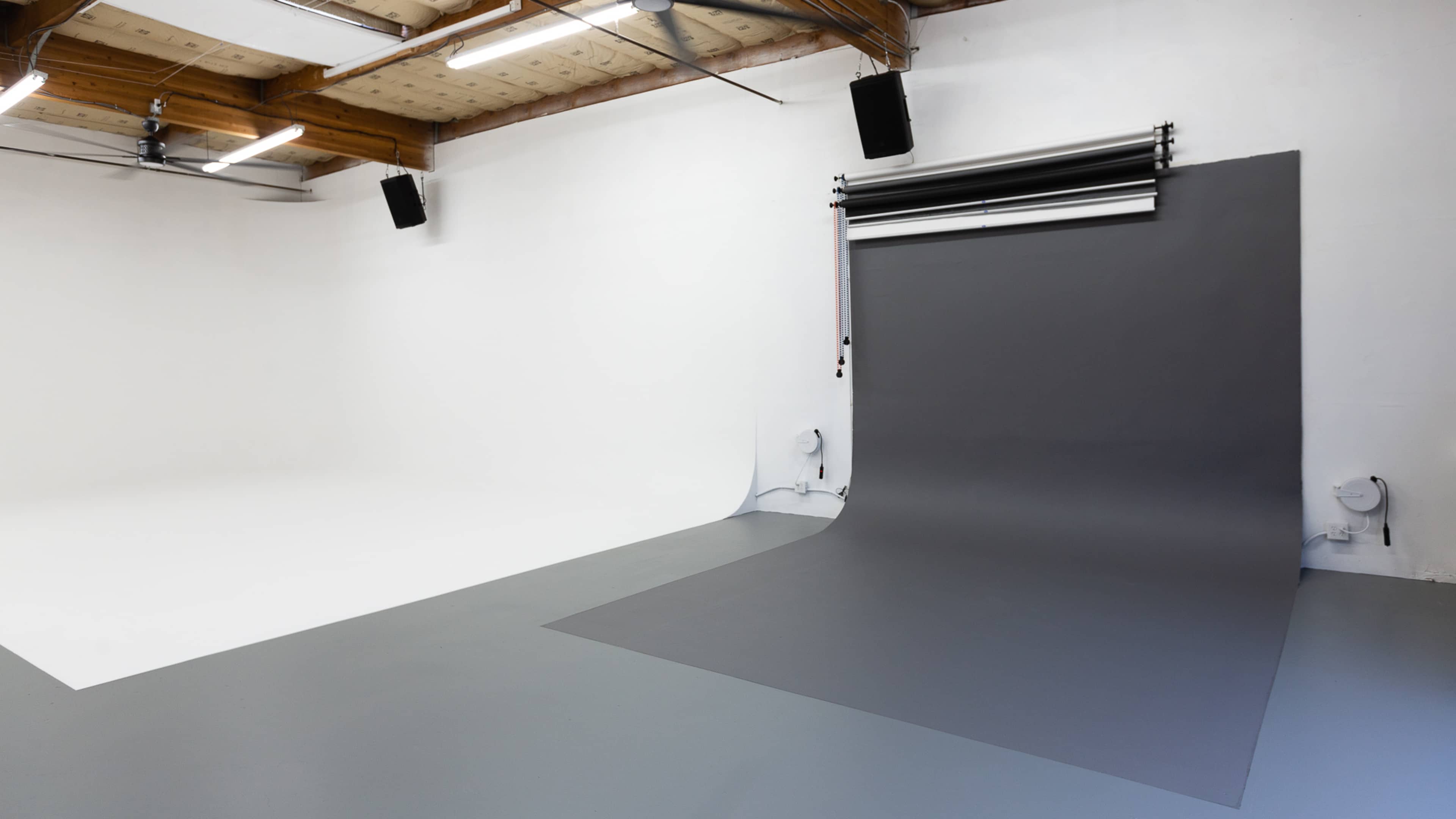 A minimalist photography studio with a white backdrop on one side and a gray backdrop on the other, featuring industrial ceiling beams and overhead lighting.