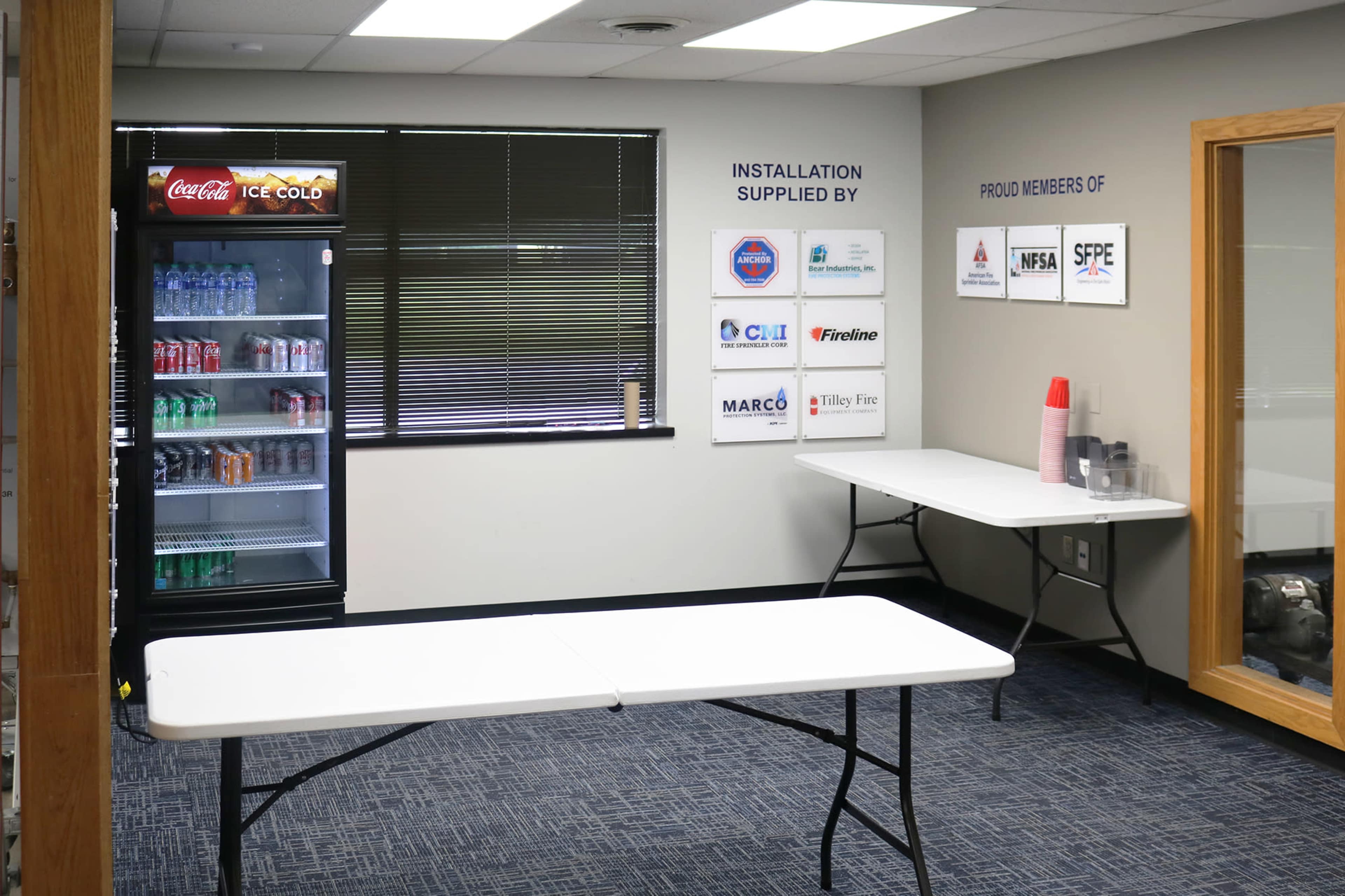 The image shows a small, sparsely furnished room with a floor of blue carpet, a vending machine filled with beverages, two white tables, and a wall displaying several membership plaques and signs.