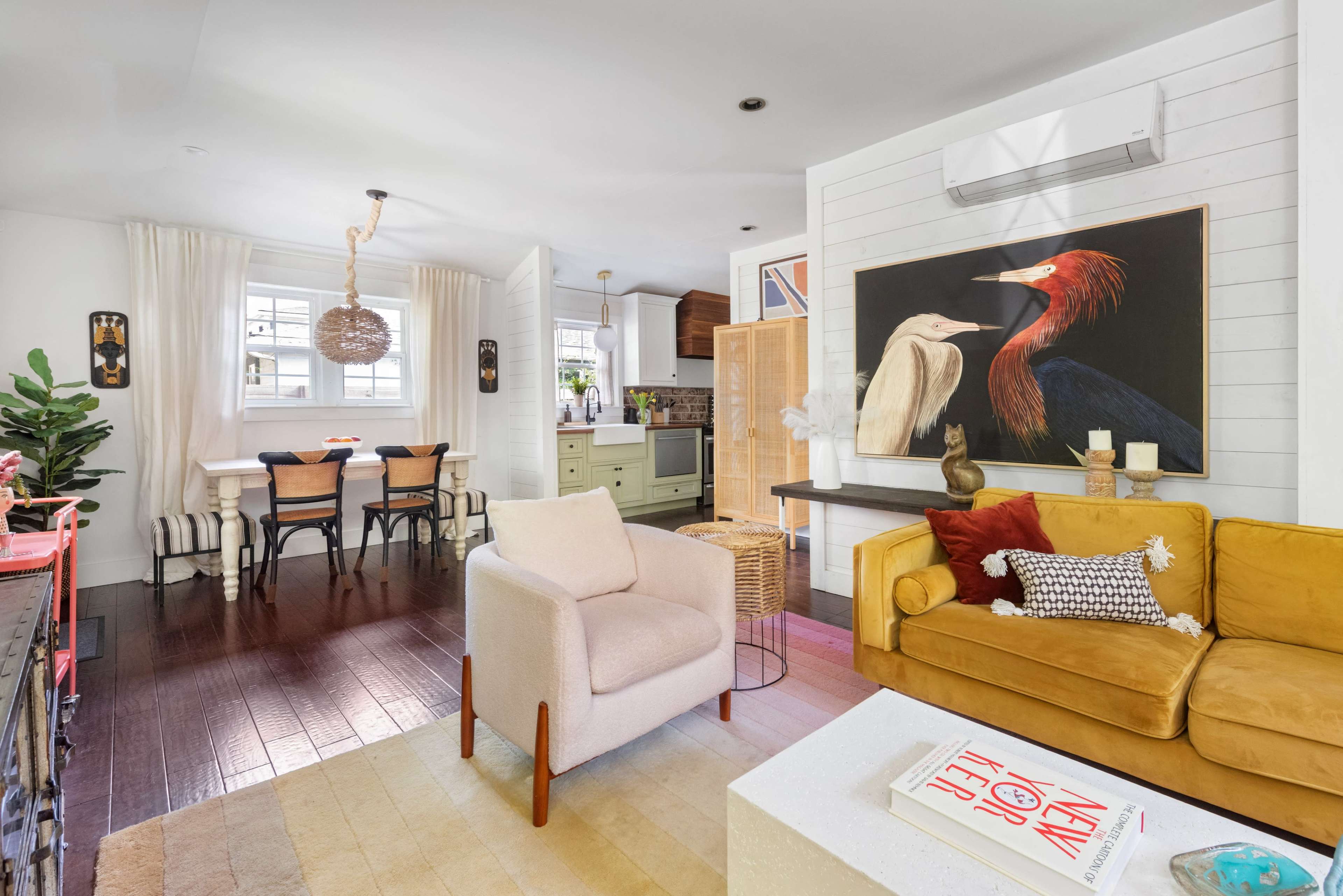 The image shows a bright living space featuring a yellow sofa, a light-colored armchair, and a dining area with a wooden table and chairs, complemented by artwork on the walls and plants.