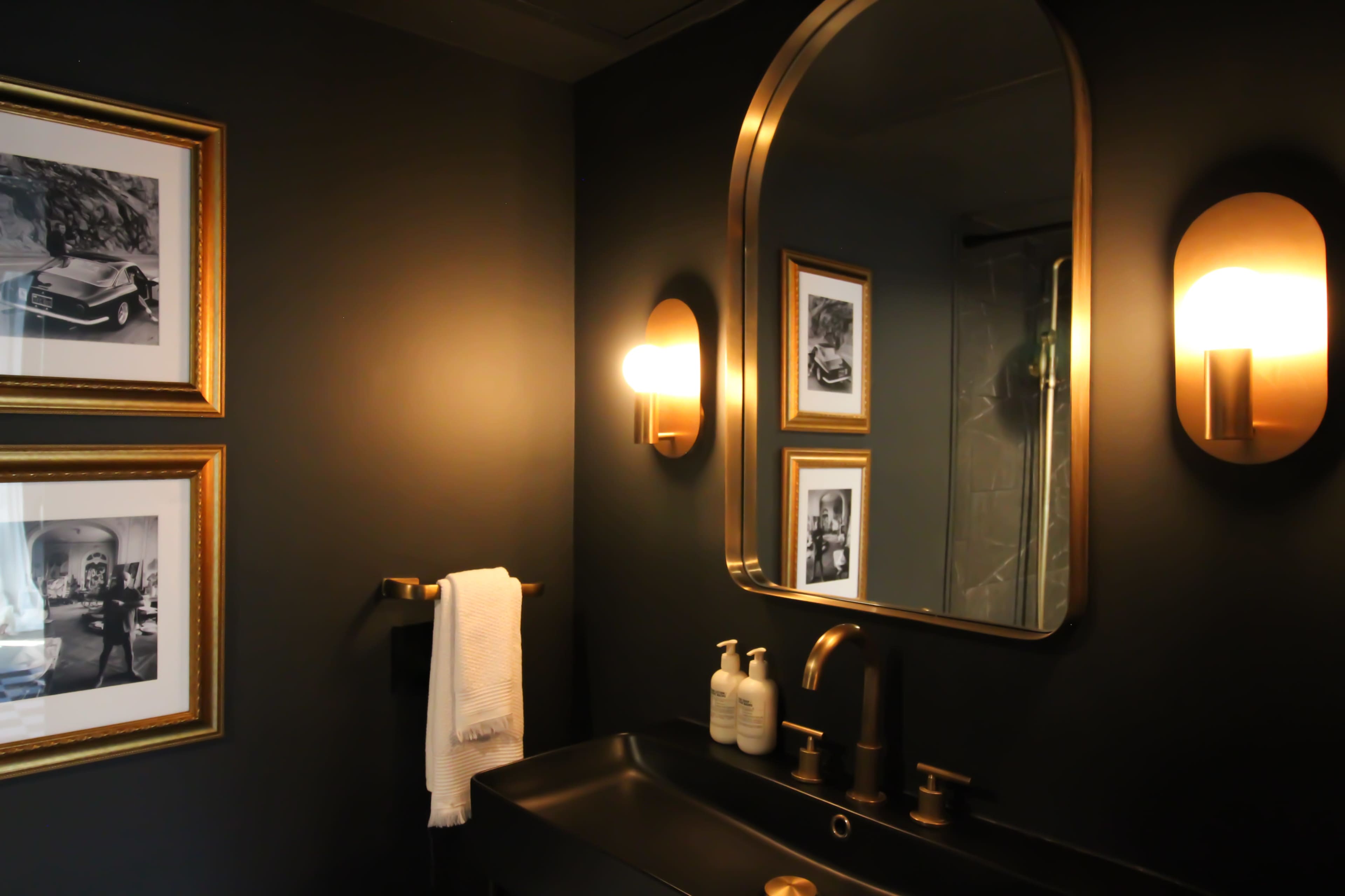 A modern bathroom with dark walls, a large mirror, gold fixtures, and framed black-and-white photographs.