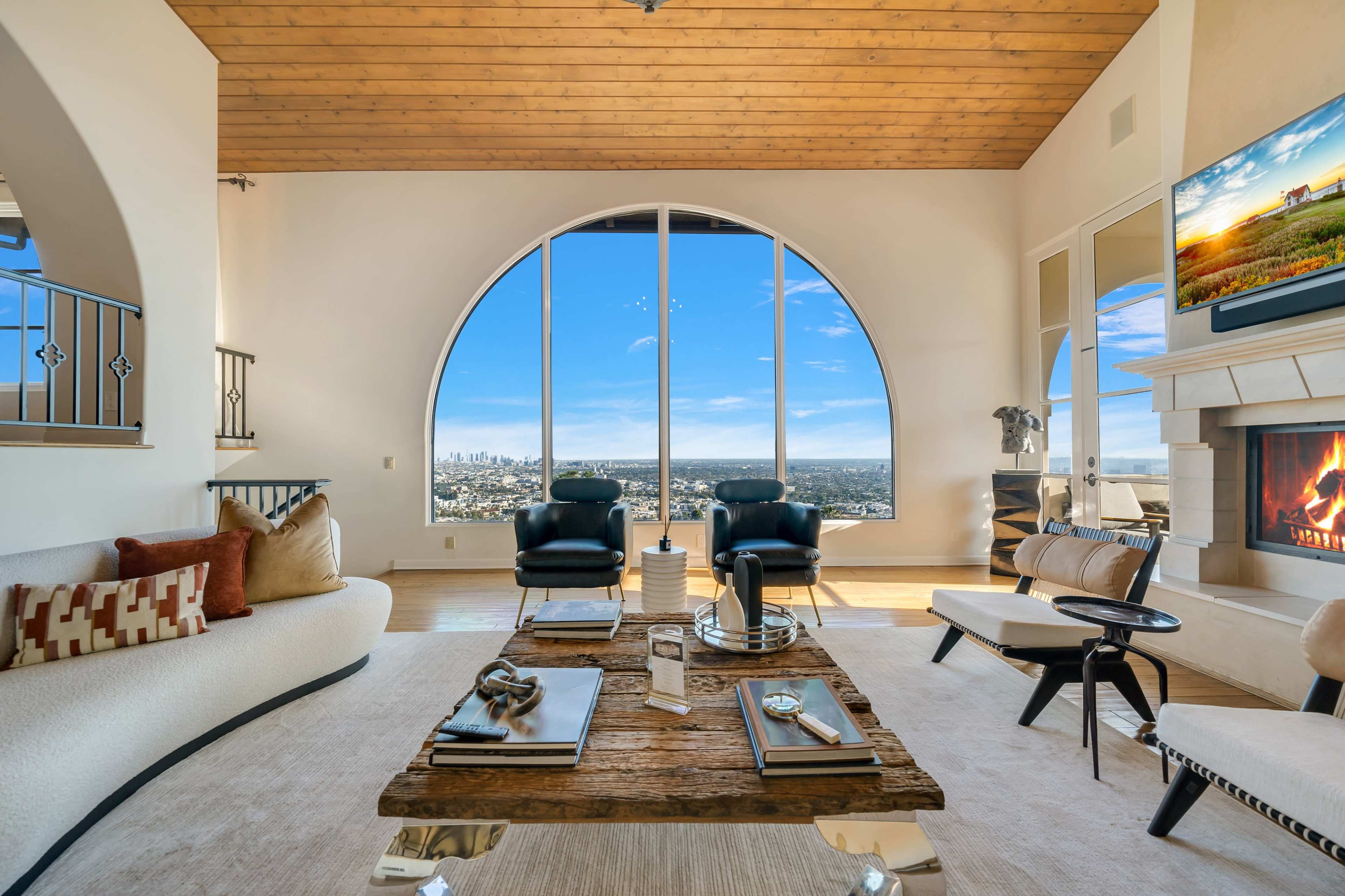 The living room features large arched windows with a city view, a wooden ceiling, a stone fireplace, and modern furniture.