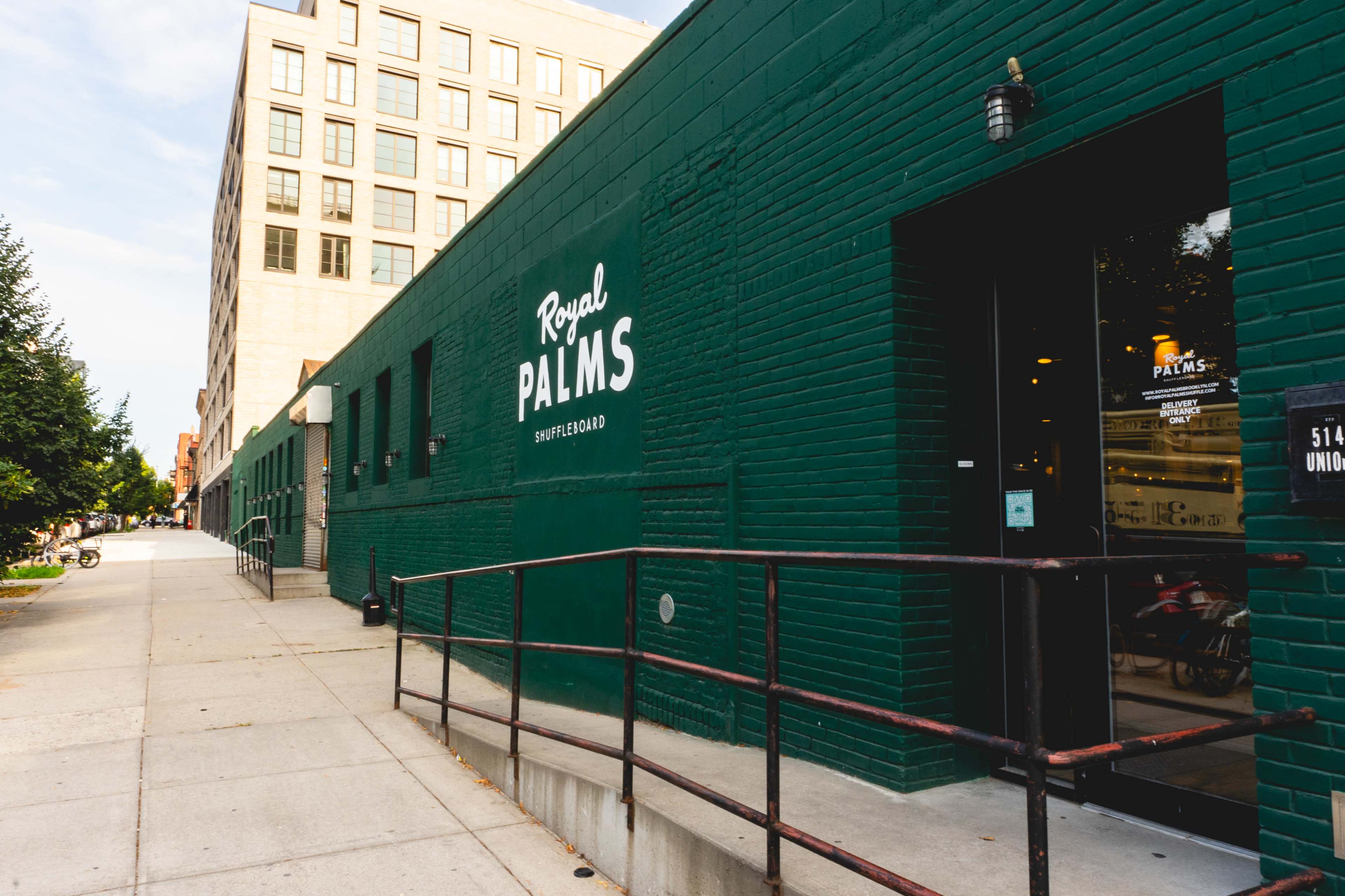 The image shows a green-brick building with the sign "Royal Palms" and a sidewalk lined with trees and buildings on either side.