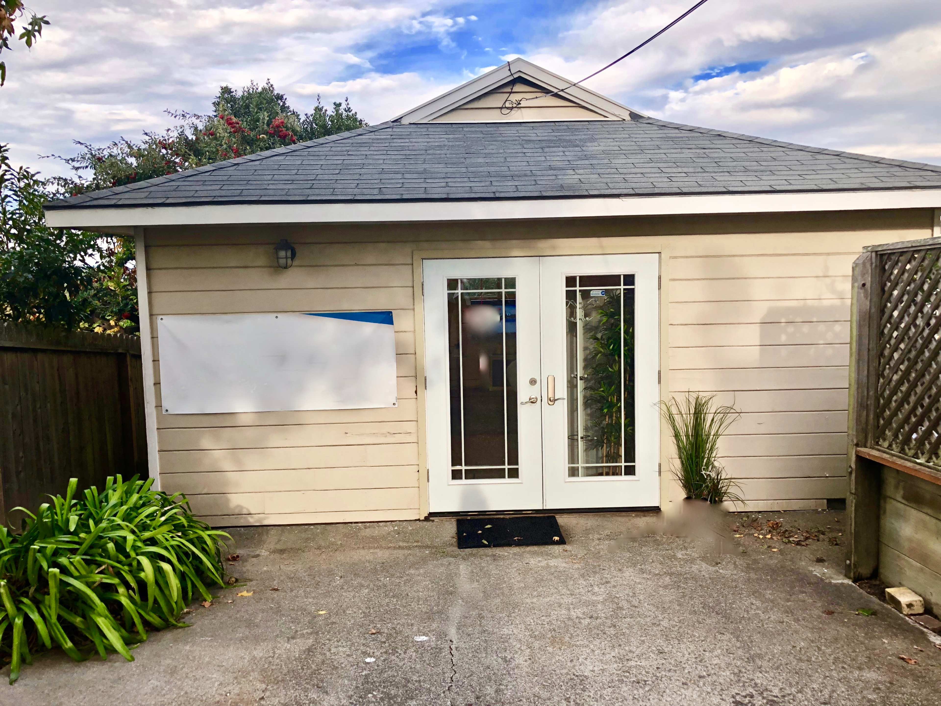 A small gray house with a gable roof, featuring double glass doors and a blank white sign on the side wall.