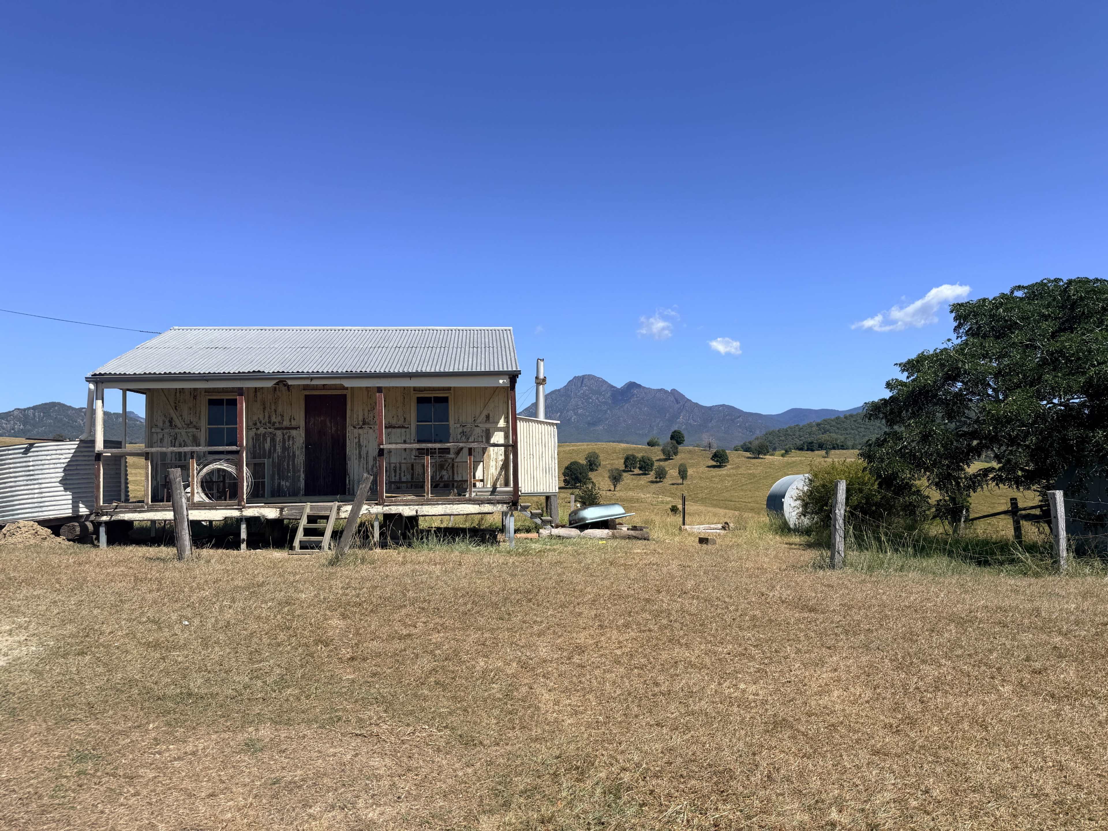 A small weathered cabin stands in a grassy field with mountains in the background under a clear blue sky.