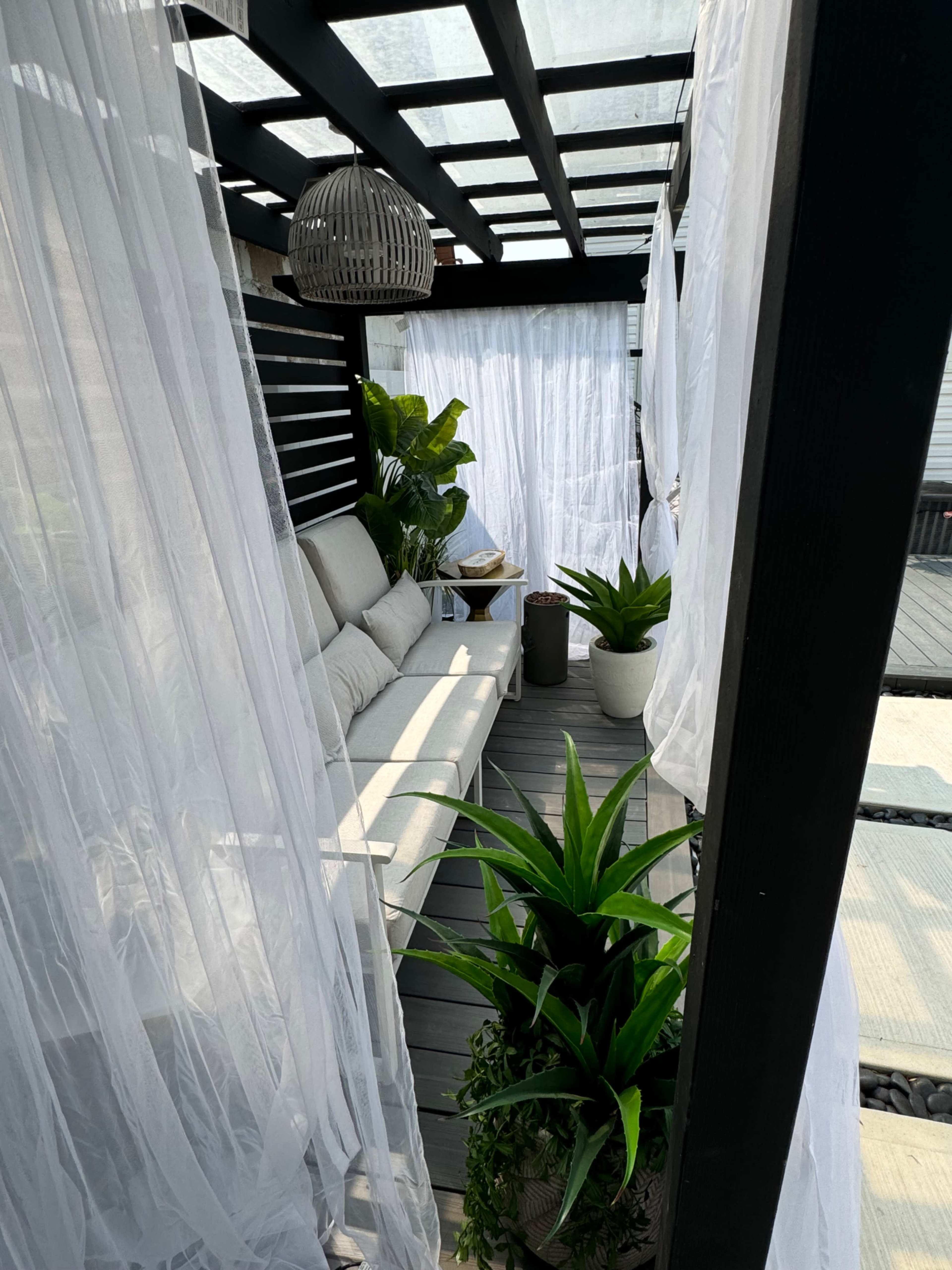 The image shows a covered outdoor area featuring a sofa, potted plants, and sheer curtains draping from a pergola.