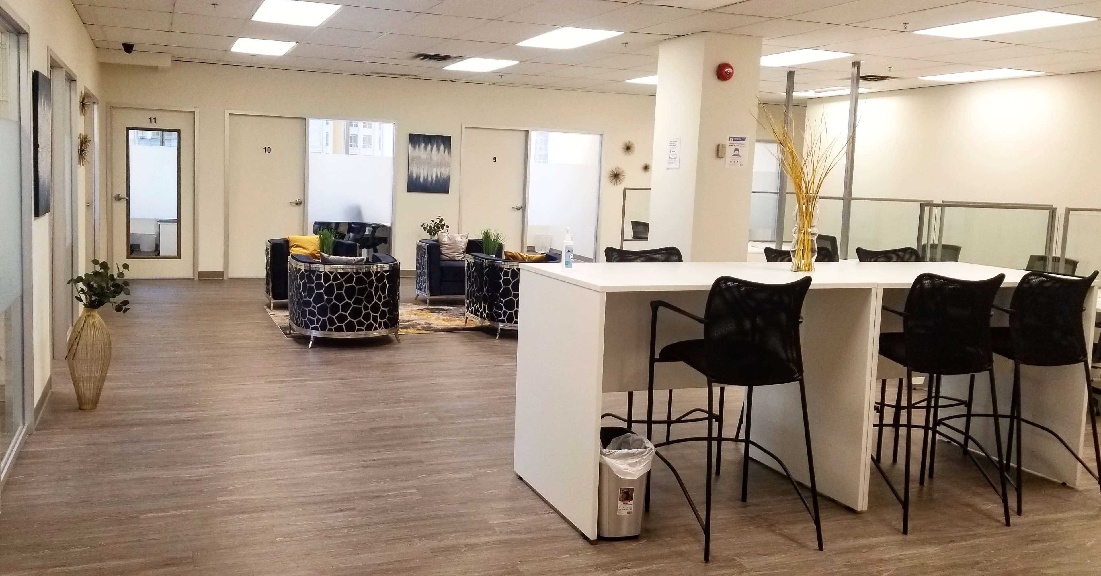 A modern office space with a kitchenette area and seating arrangements, featuring black and yellow chairs, a coffee table, and plants.