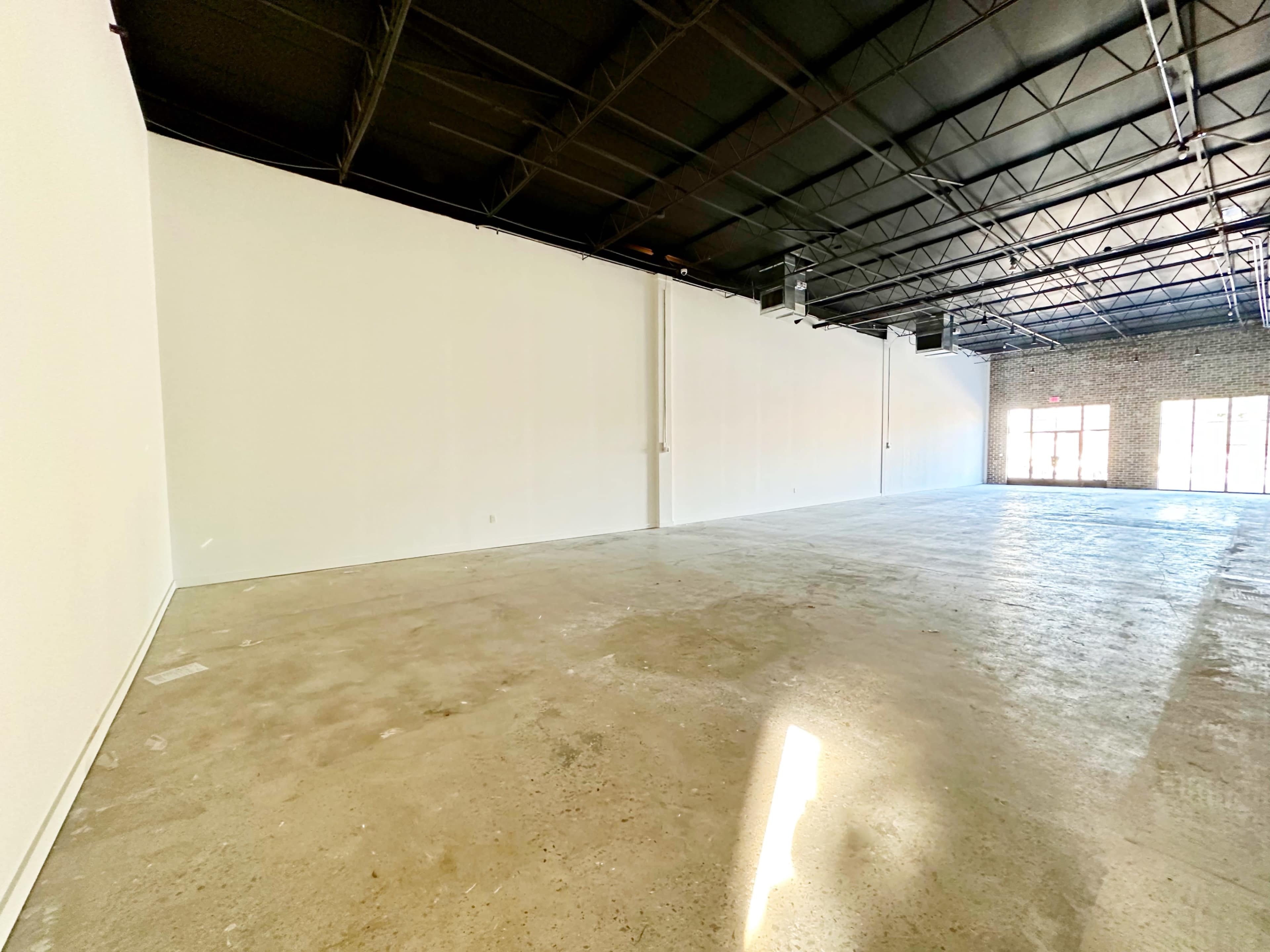 An empty commercial space features concrete flooring and white walls, with large windows on one side allowing natural light to enter.