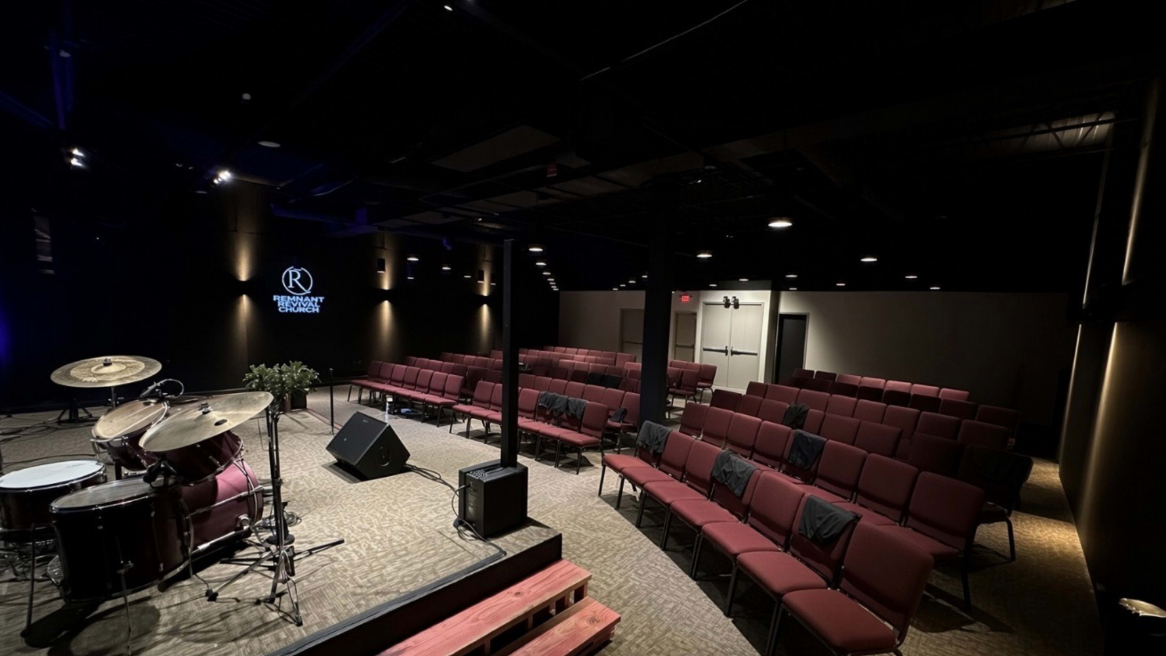 The image shows an indoor performance space with rows of maroon chairs arranged in front of a small stage with a drum set.