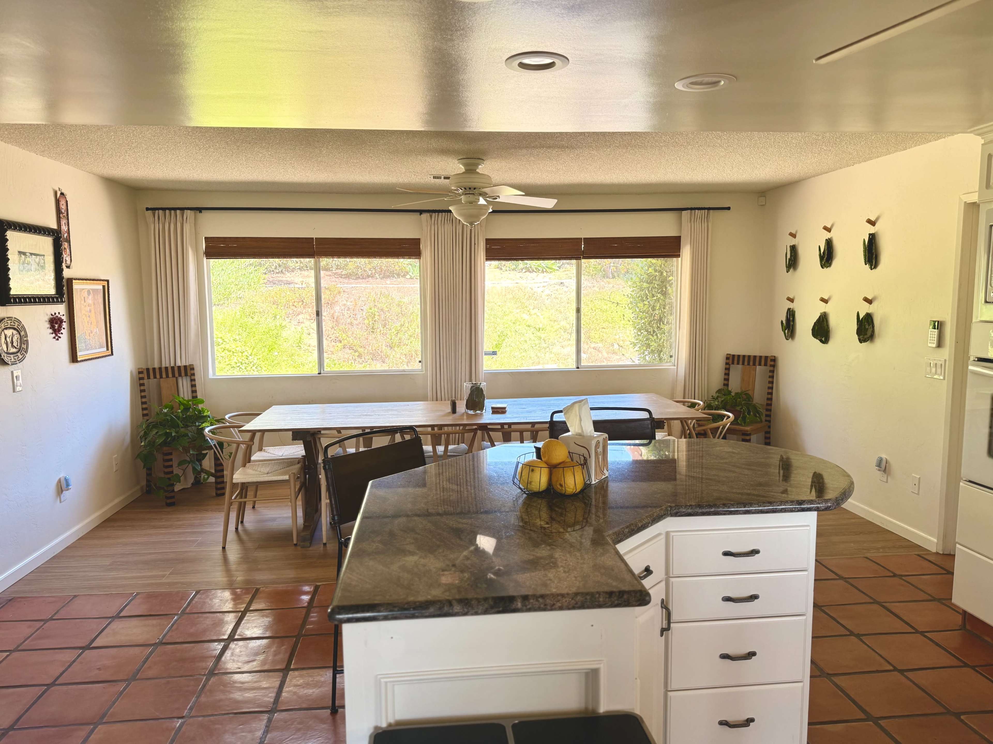 The image shows a bright, open kitchen and dining area featuring a central island, a wooden dining table, and large windows with natural light streaming in.