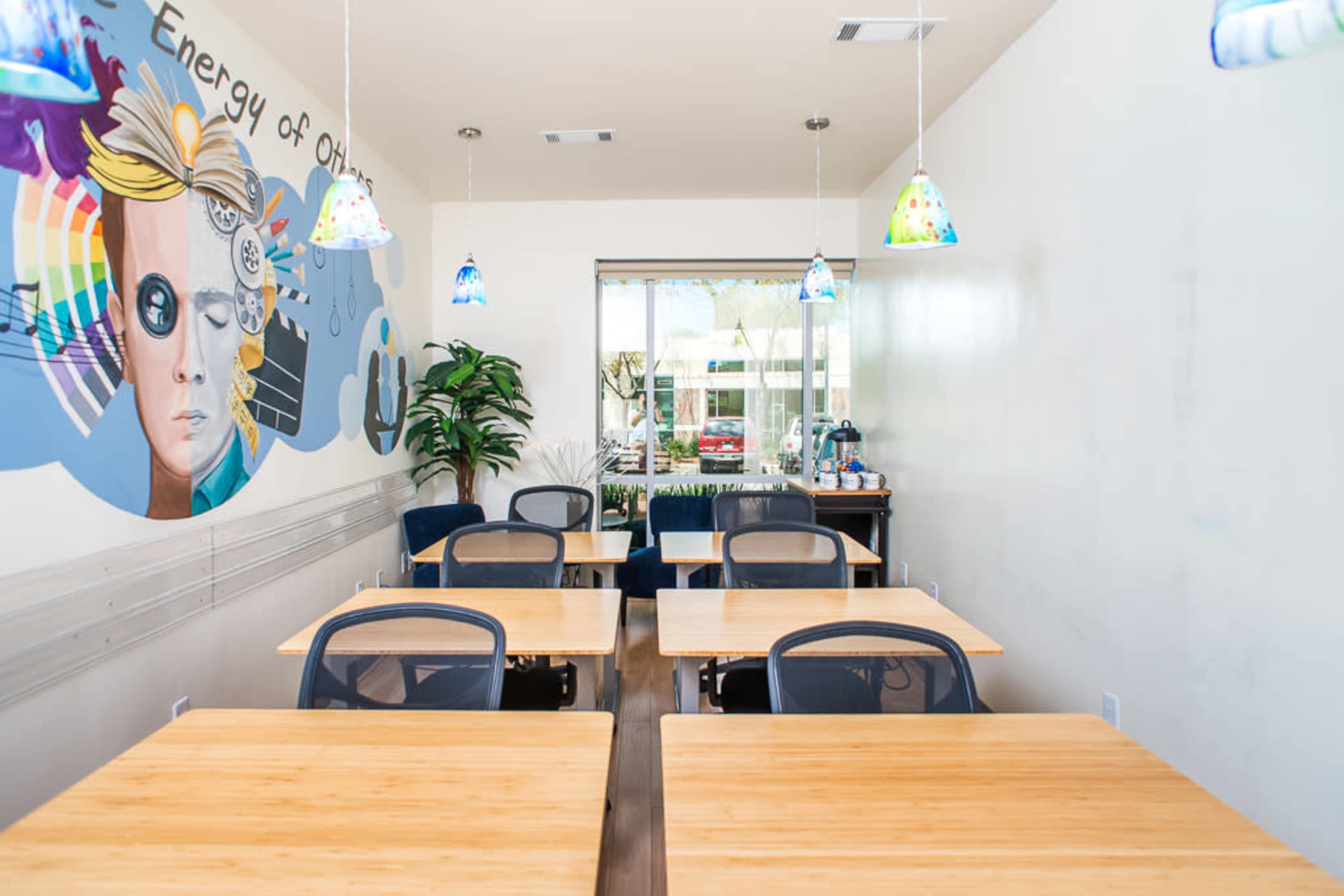 A bright dining area features wooden tables and chairs, with a mural on the wall and colorful pendant lights hanging above.