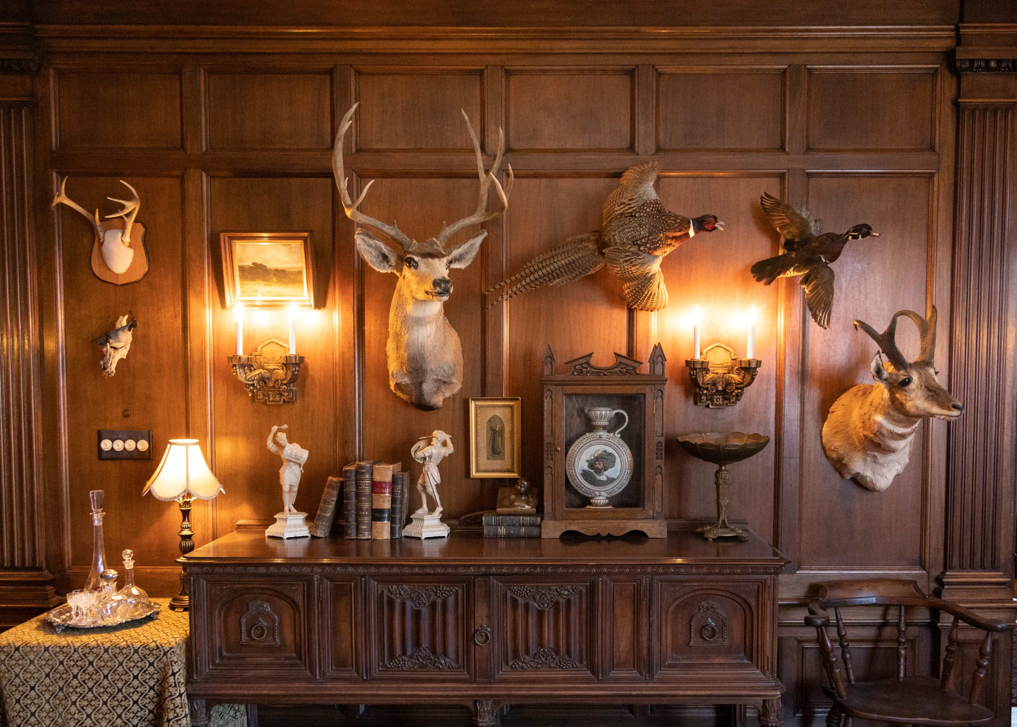 The image shows a wooden wall adorned with mounted animal heads and a vintage clock, accompanied by a side table featuring decorative items and books.