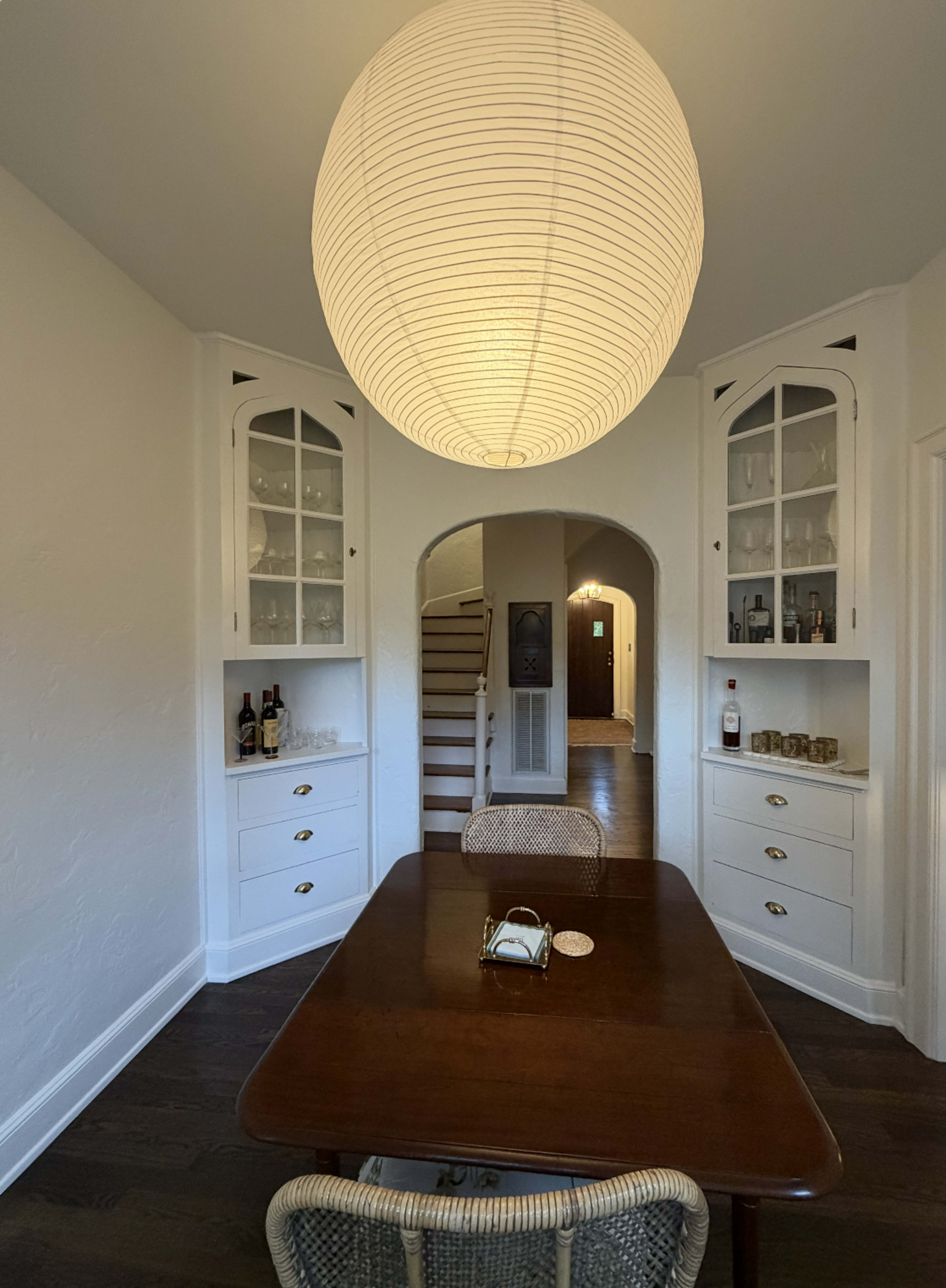 The image shows a dining area with a wooden table, a large round pendant light, and built-in cabinets on either side, leading to a hallway and staircase in the background.