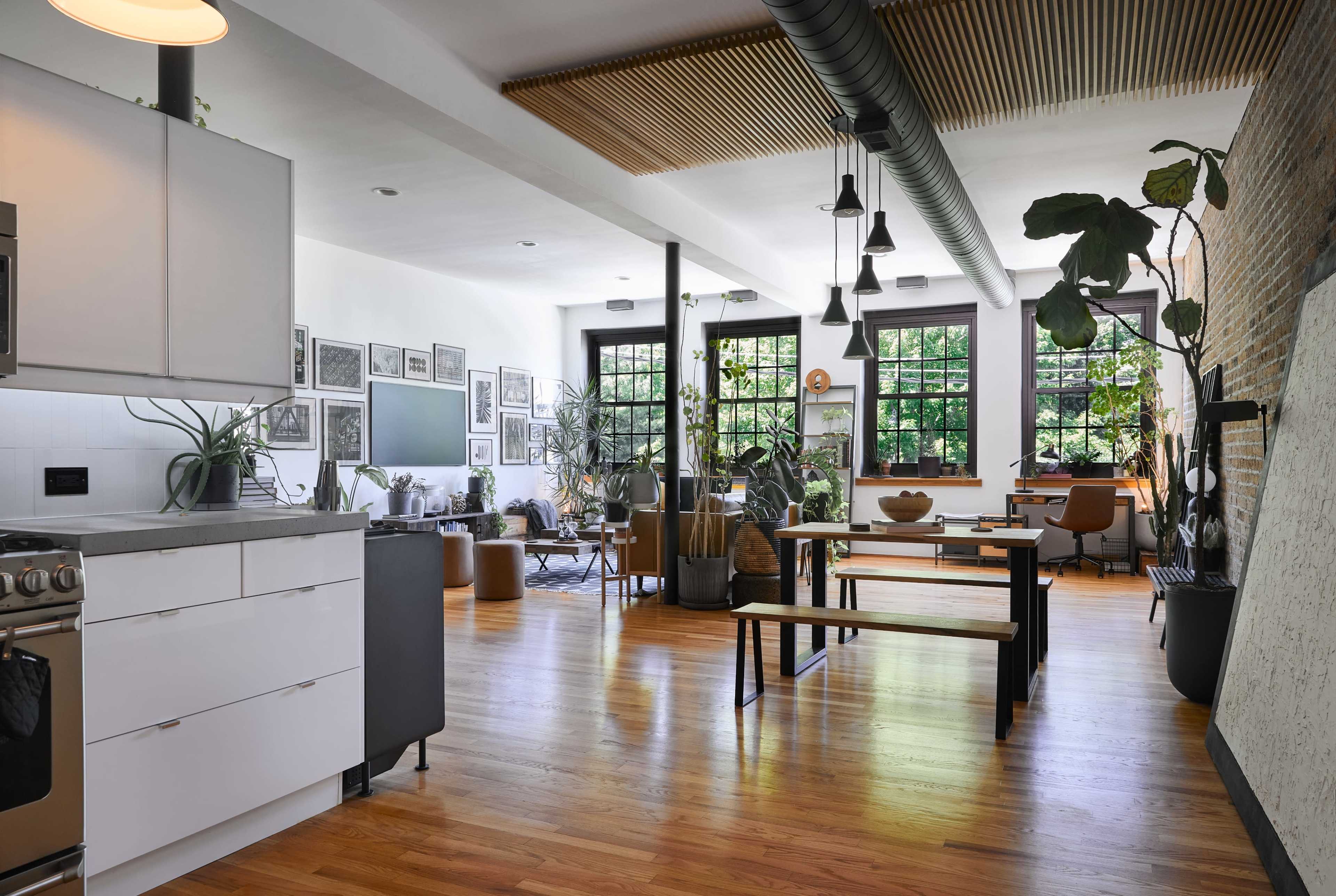 The image depicts a modern, open-concept living space with a kitchen, dining area, and seating area, featuring wooden flooring and numerous plants throughout.