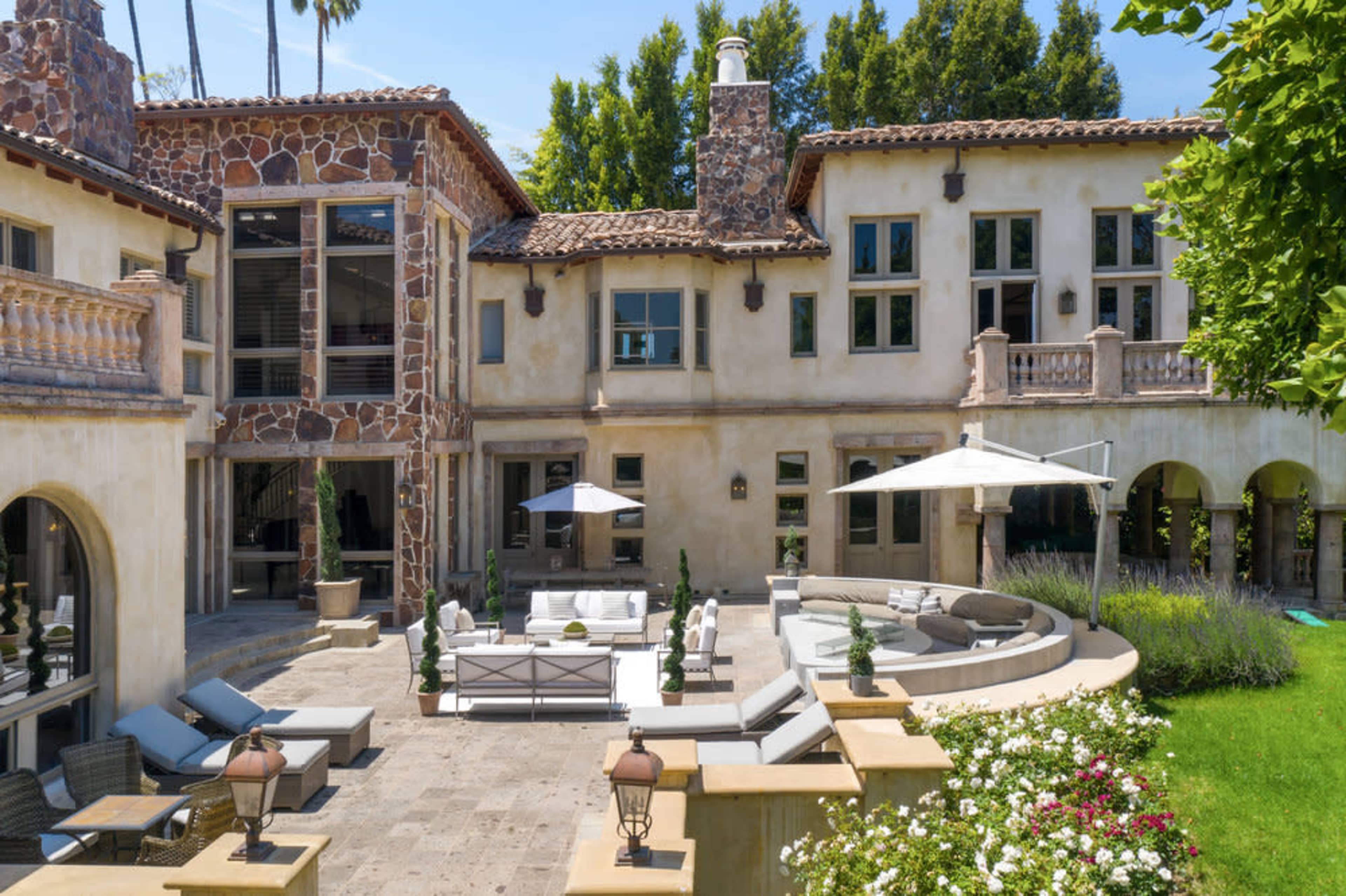 The image shows a spacious outdoor patio area of a luxurious home, featuring comfortable seating arrangements, an umbrella, and well-manicured landscaping.