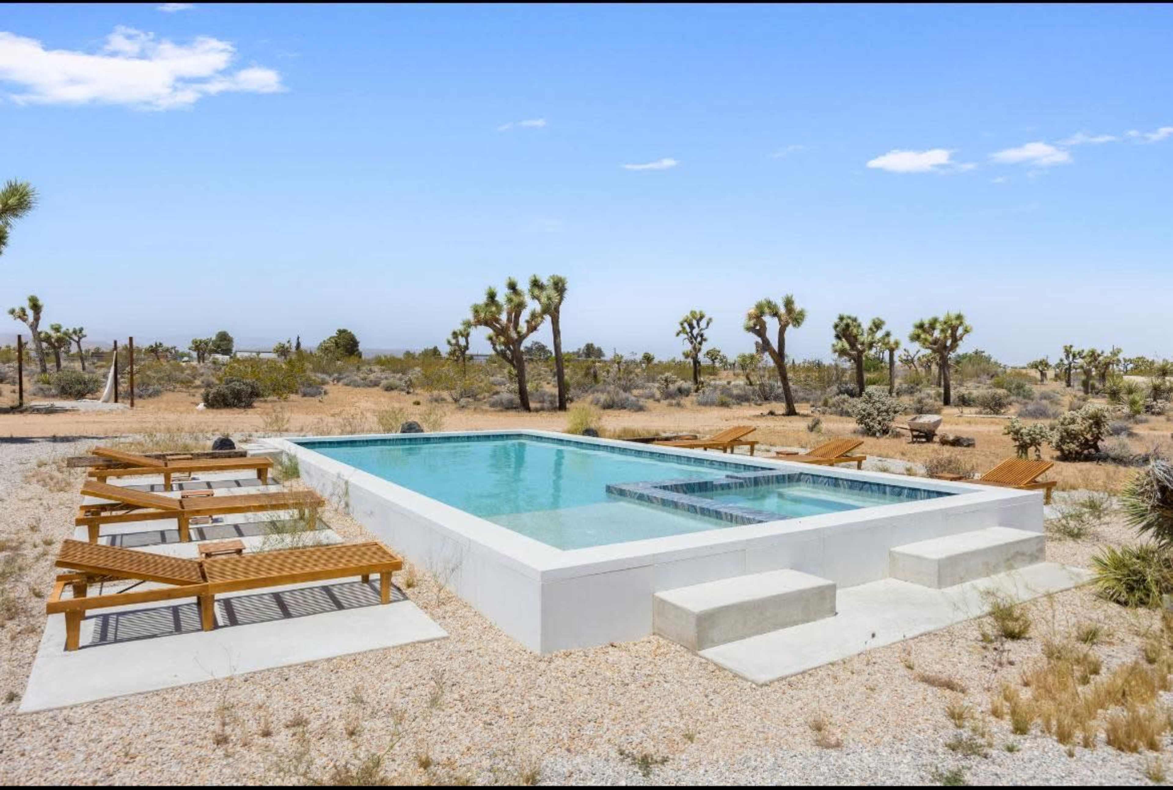 A rectangular swimming pool surrounded by wooden lounge chairs in a desert landscape with sparse vegetation and Joshua trees.