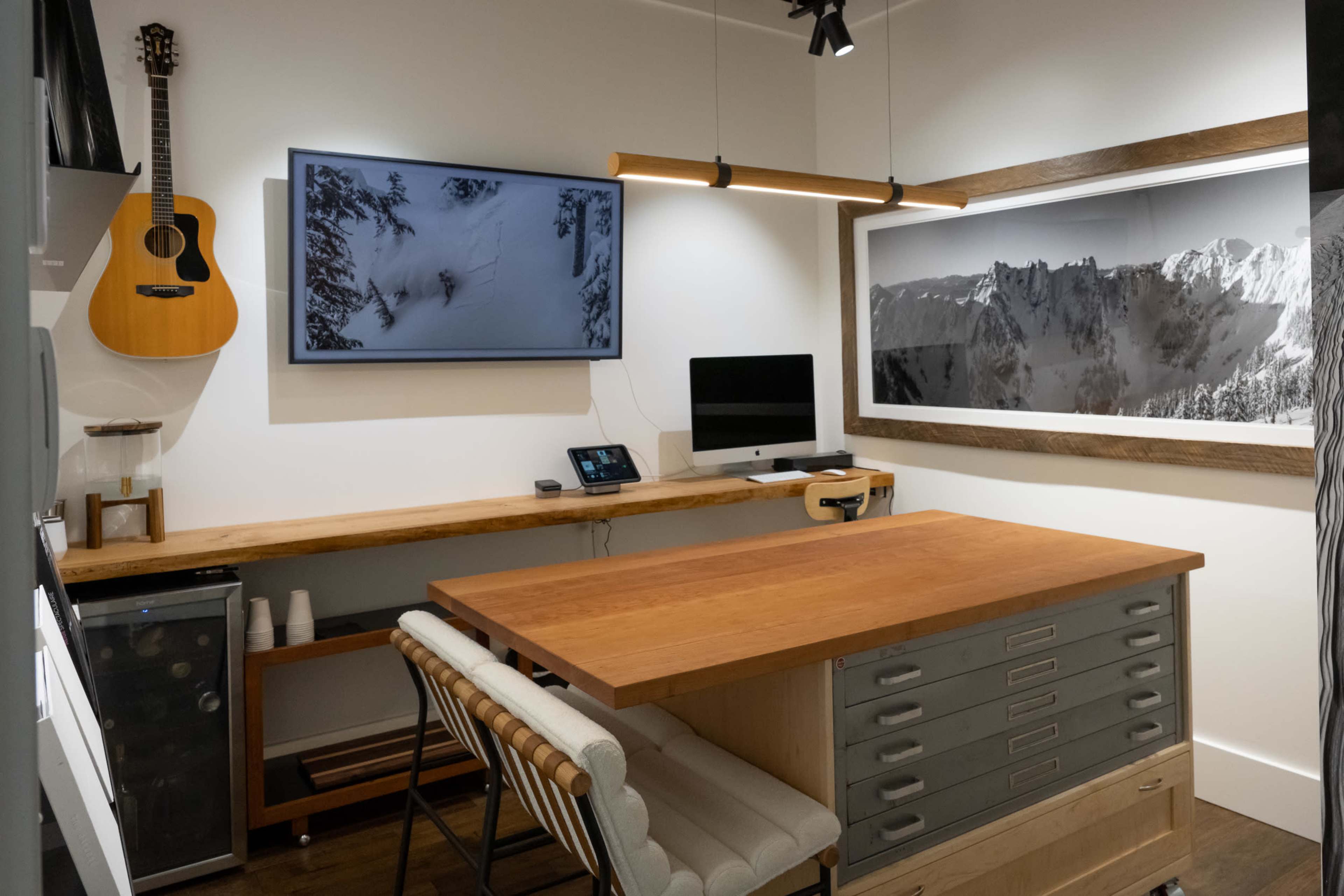 A modern workspace with a wooden table, two monitors, an acoustic guitar hanging on the wall, and a large photograph of mountains.