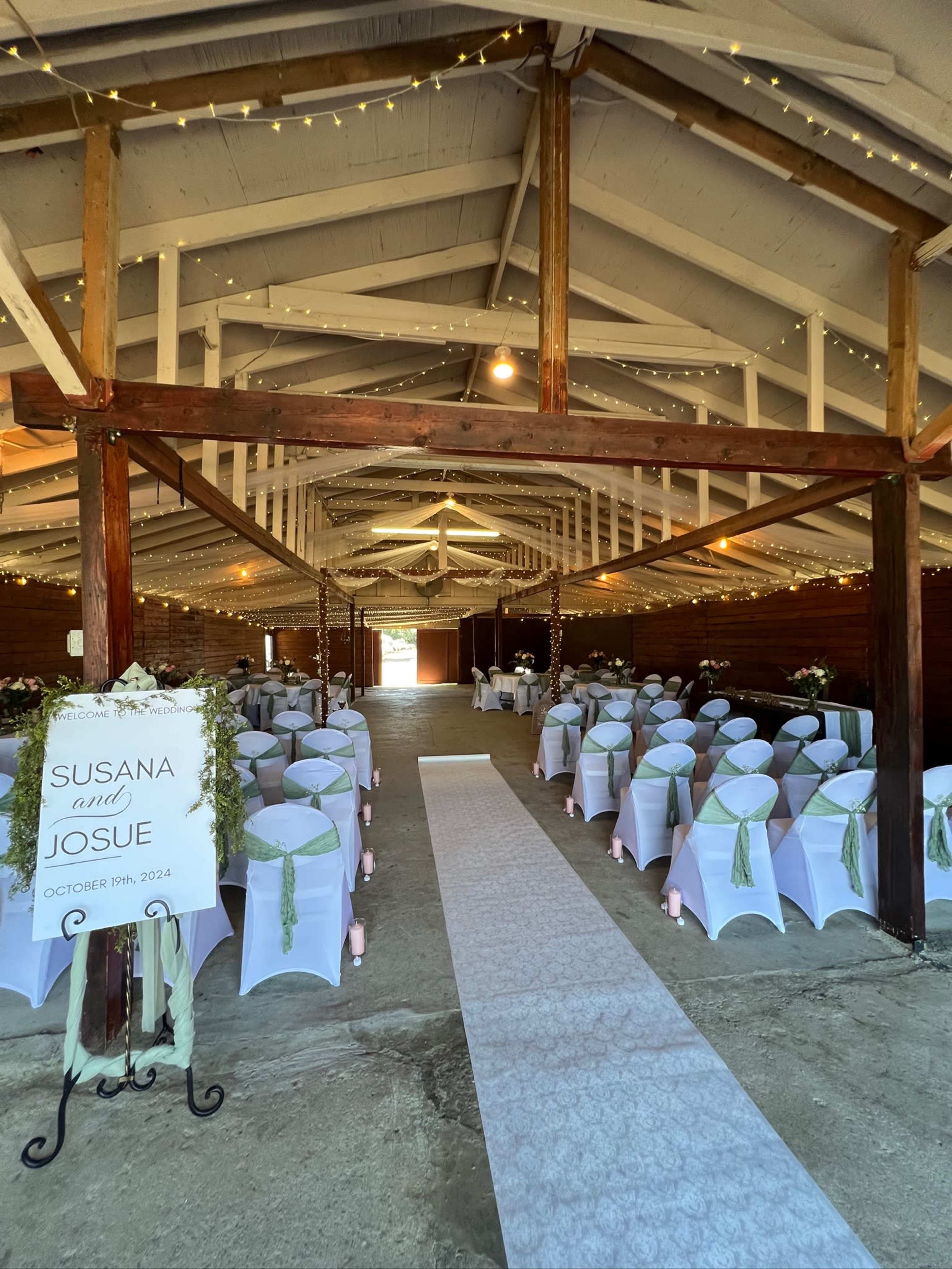 The image shows a rustic event space set up for a wedding, featuring rows of decorated tables with white chair covers and a central aisle leading to an entrance adorned with string lights.
