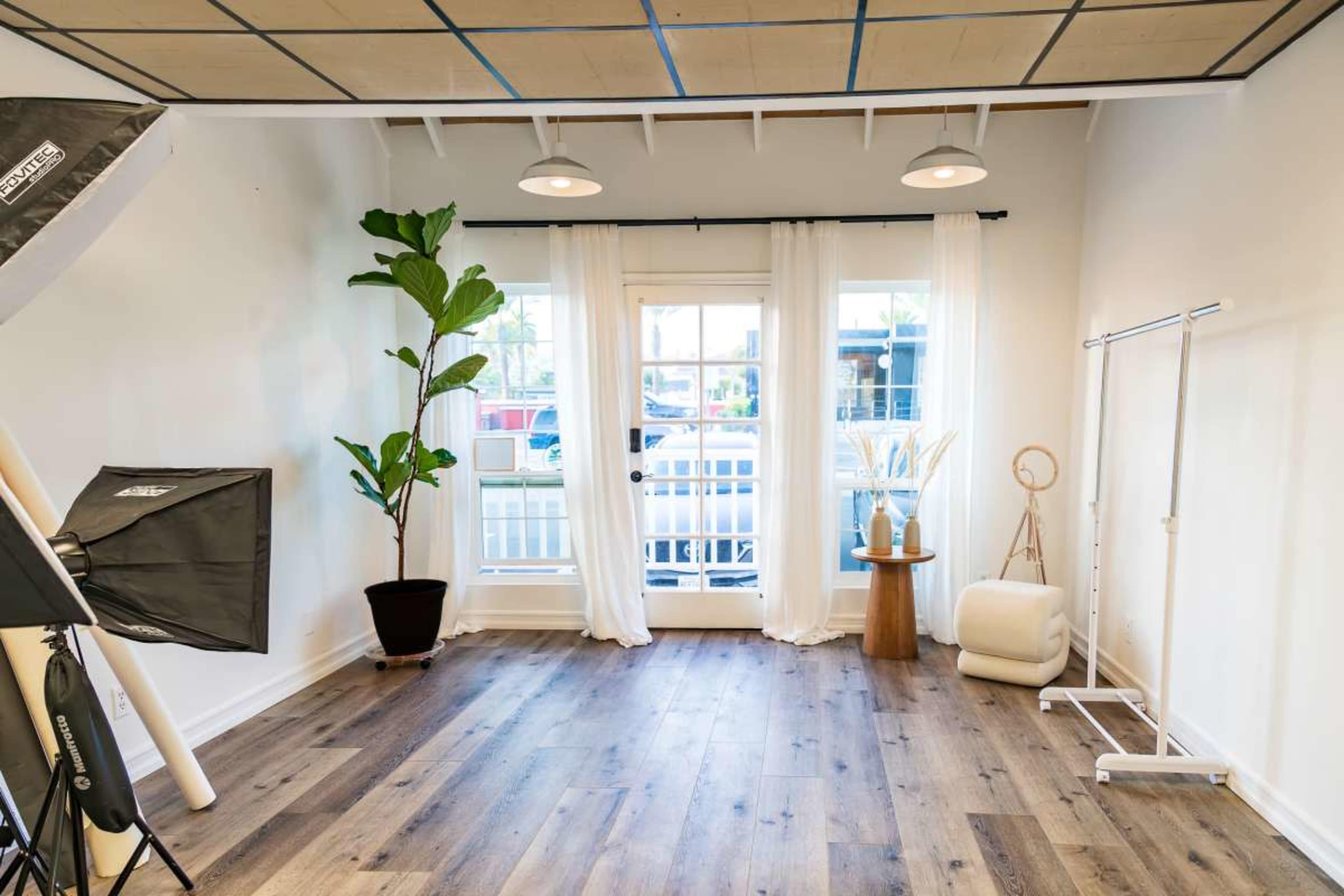 The image shows a bright, minimalist room with large windows, a potted plant, and simple furnishings including a wooden stool and a clothing rack.