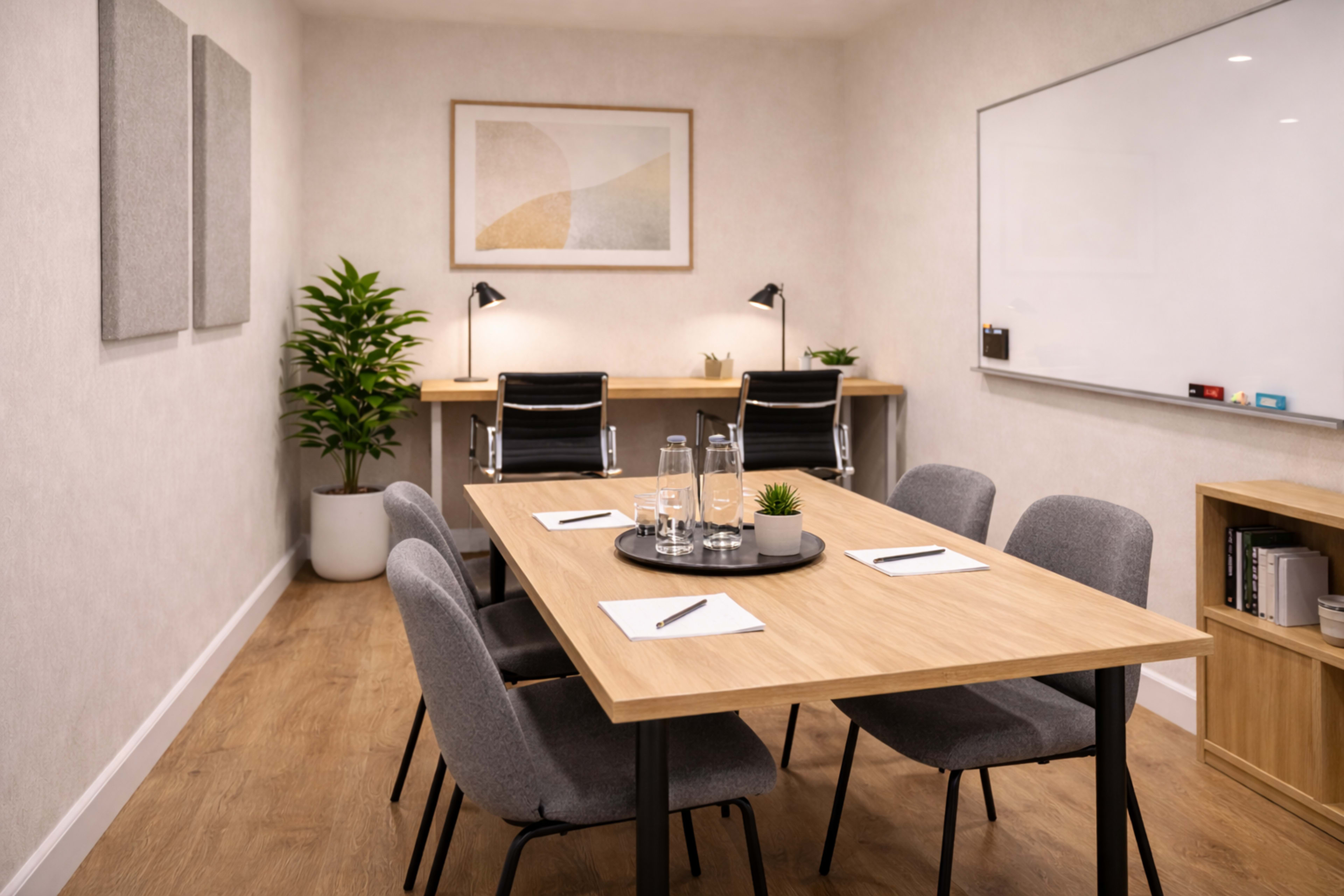 The image shows a modern meeting room featuring a wooden table surrounded by gray chairs, a whiteboard on the wall, and two desks with lamps in the background.