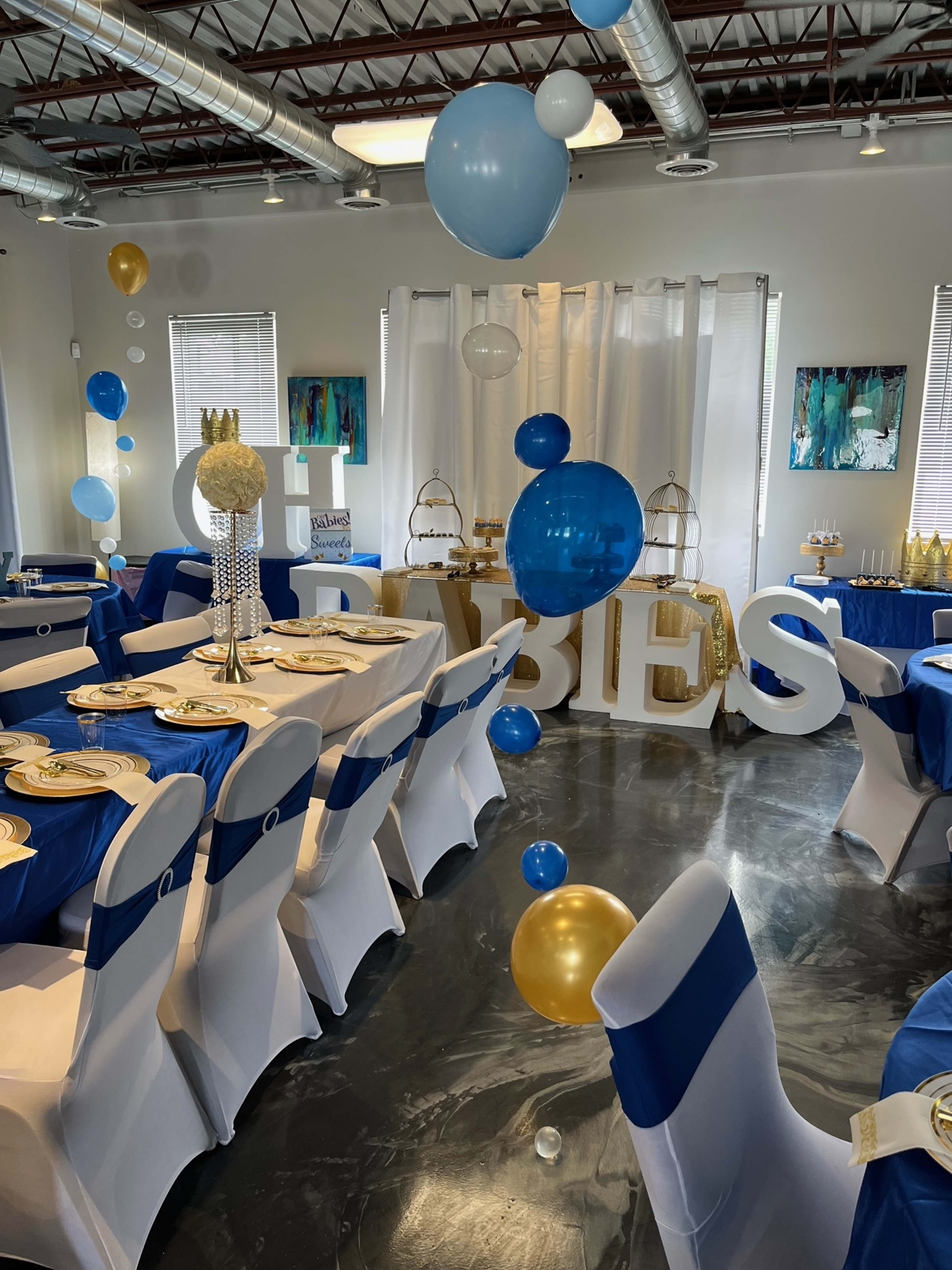 The image shows a decorated event space with tables set for a celebration, featuring blue and gold balloons, and the word "BABIES" displayed prominently in large letters.