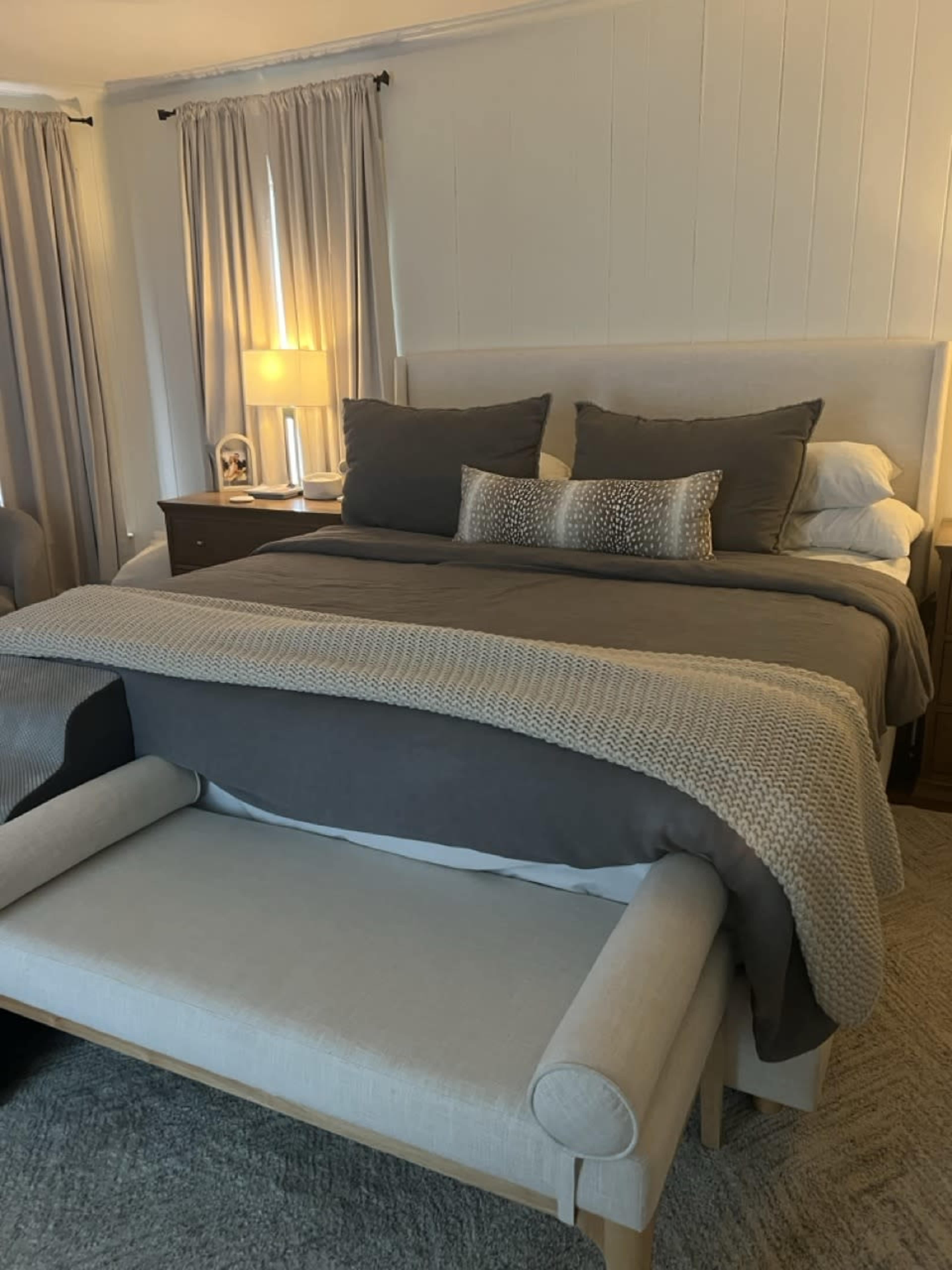 The image shows a neatly made bed with gray and white bedding, positioned in a softly lit bedroom with curtains and a lamp on a bedside table.