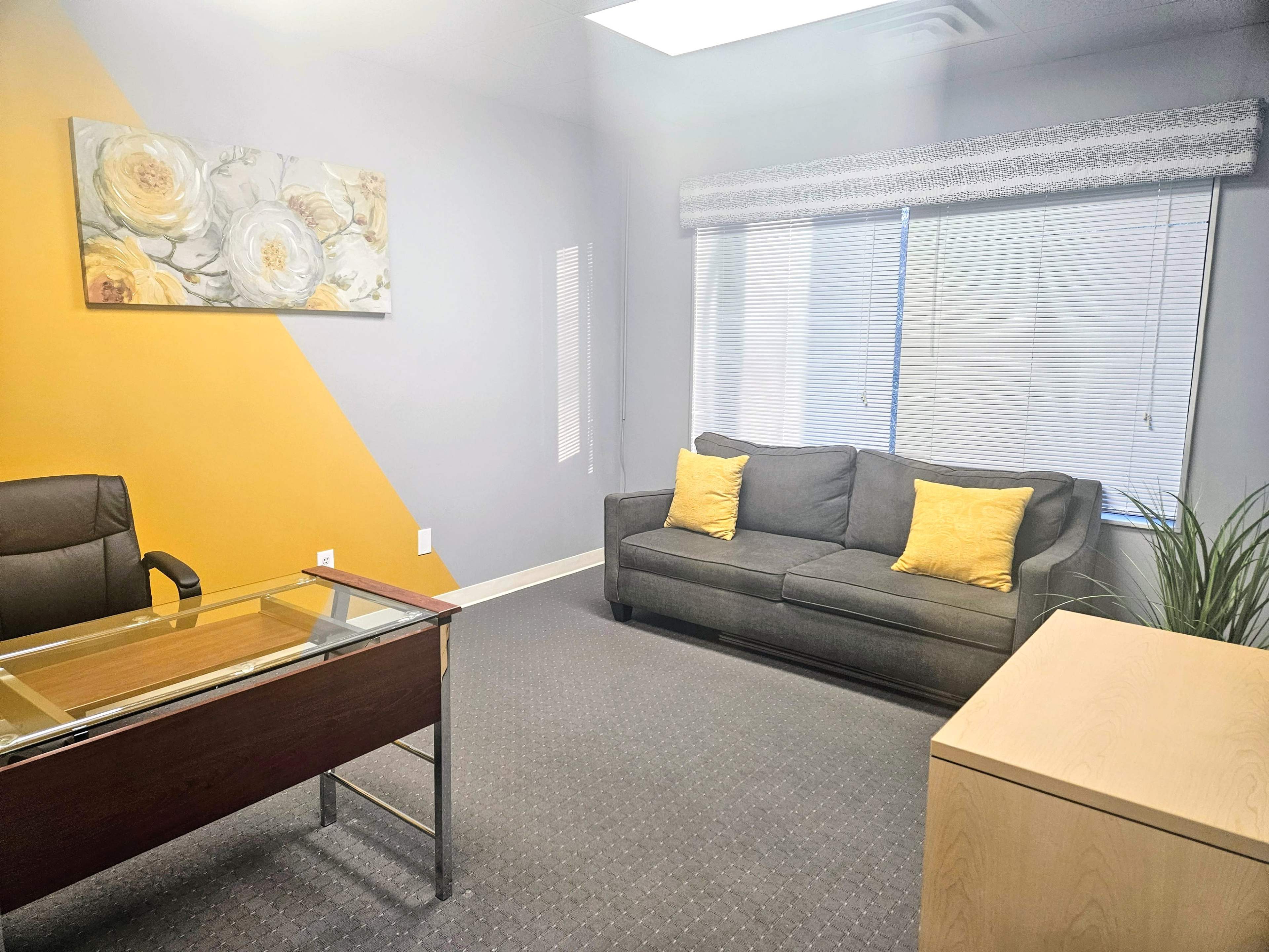 An office space with a glass-top desk, a gray couch with yellow pillows, and large windows with blinds.