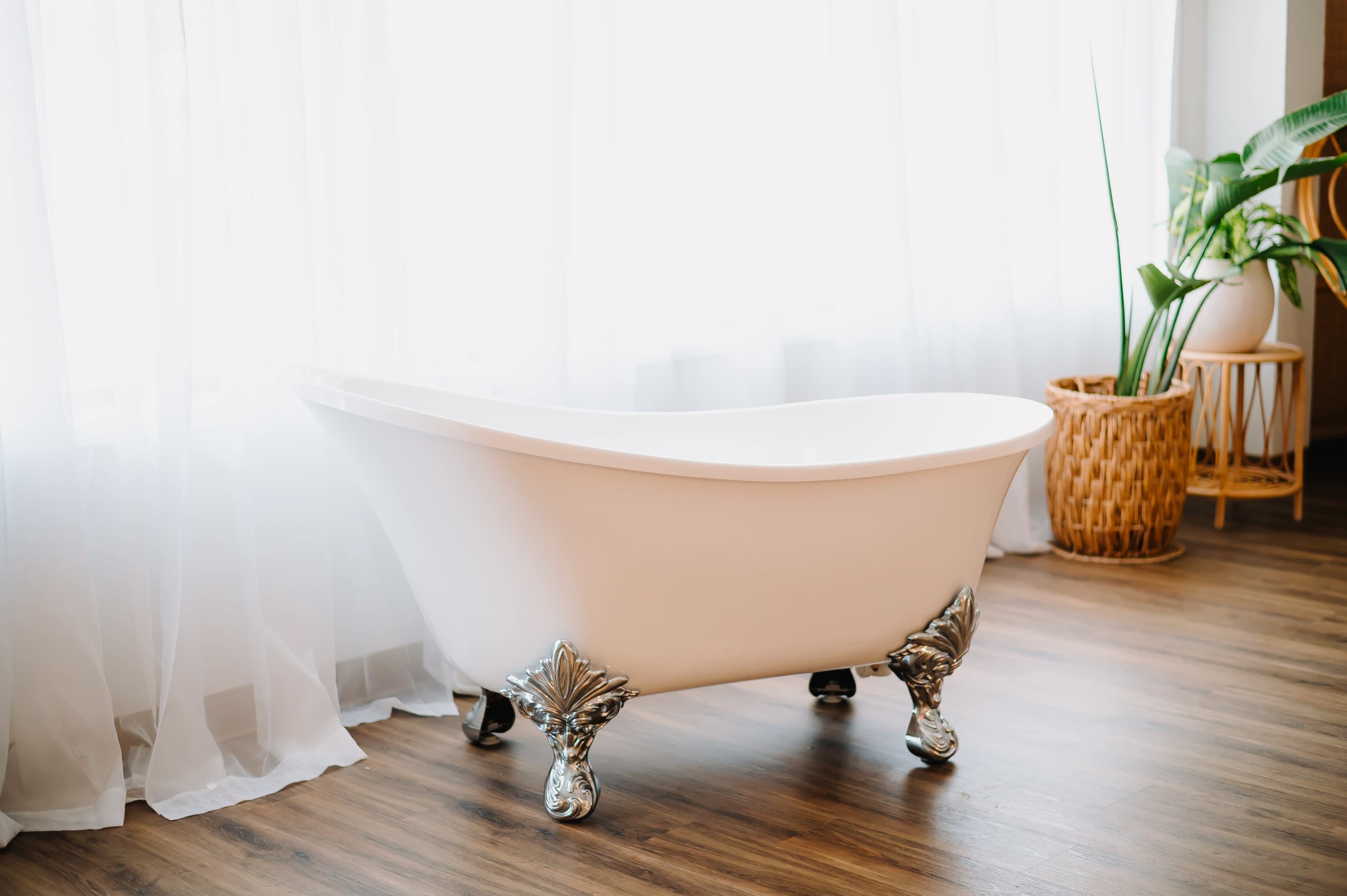 A freestanding white bathtub with decorative silver feet is positioned beside a window with sheer curtains.