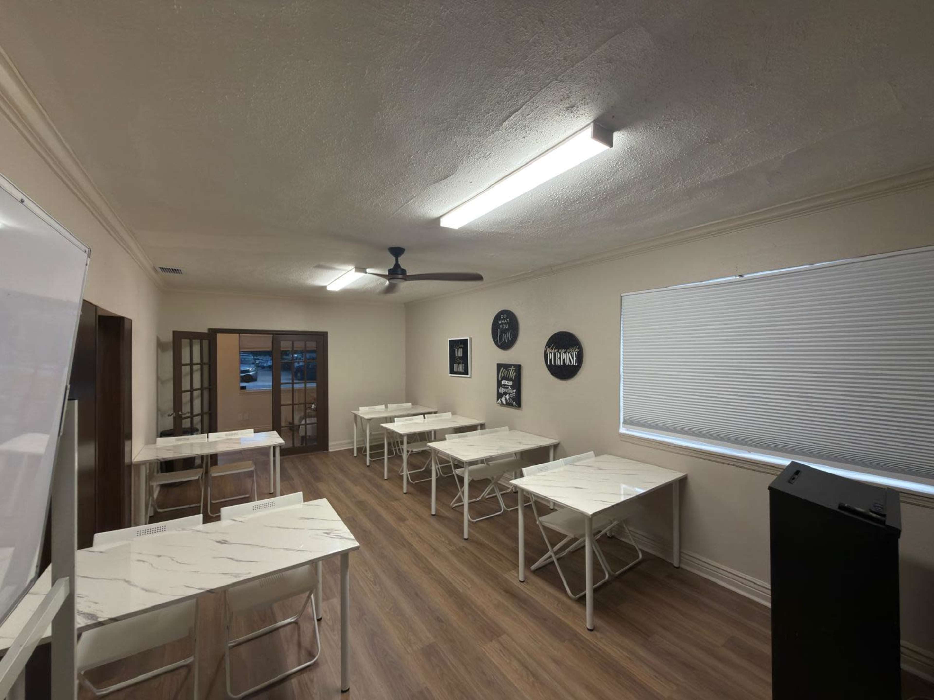 A well-lit classroom features several white tables arranged neatly with a fan on the ceiling and a window with blinds.