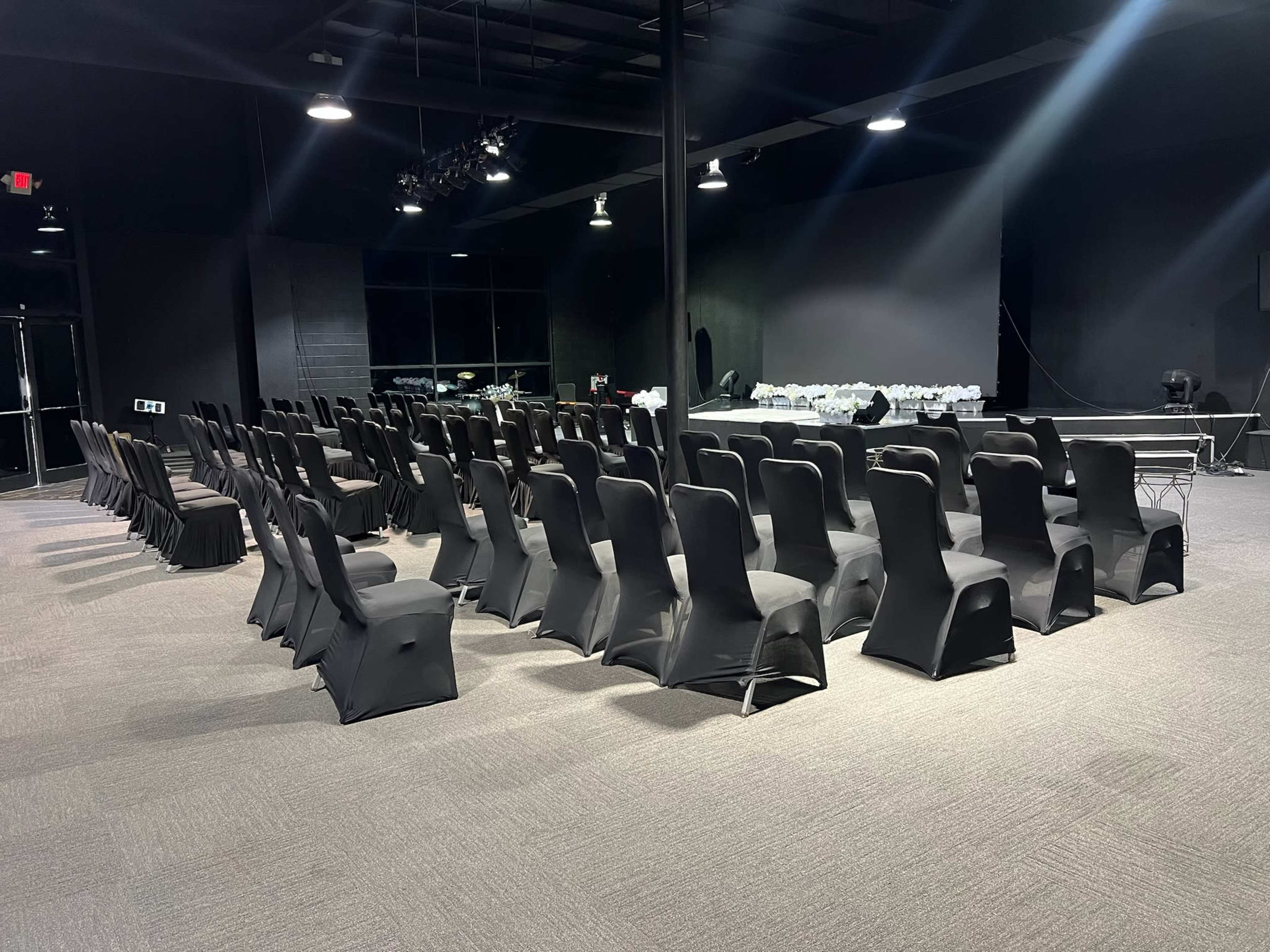 The image shows a large room set up for an event, featuring rows of black chairs arranged in a neat pattern with a stage or platform in the background.