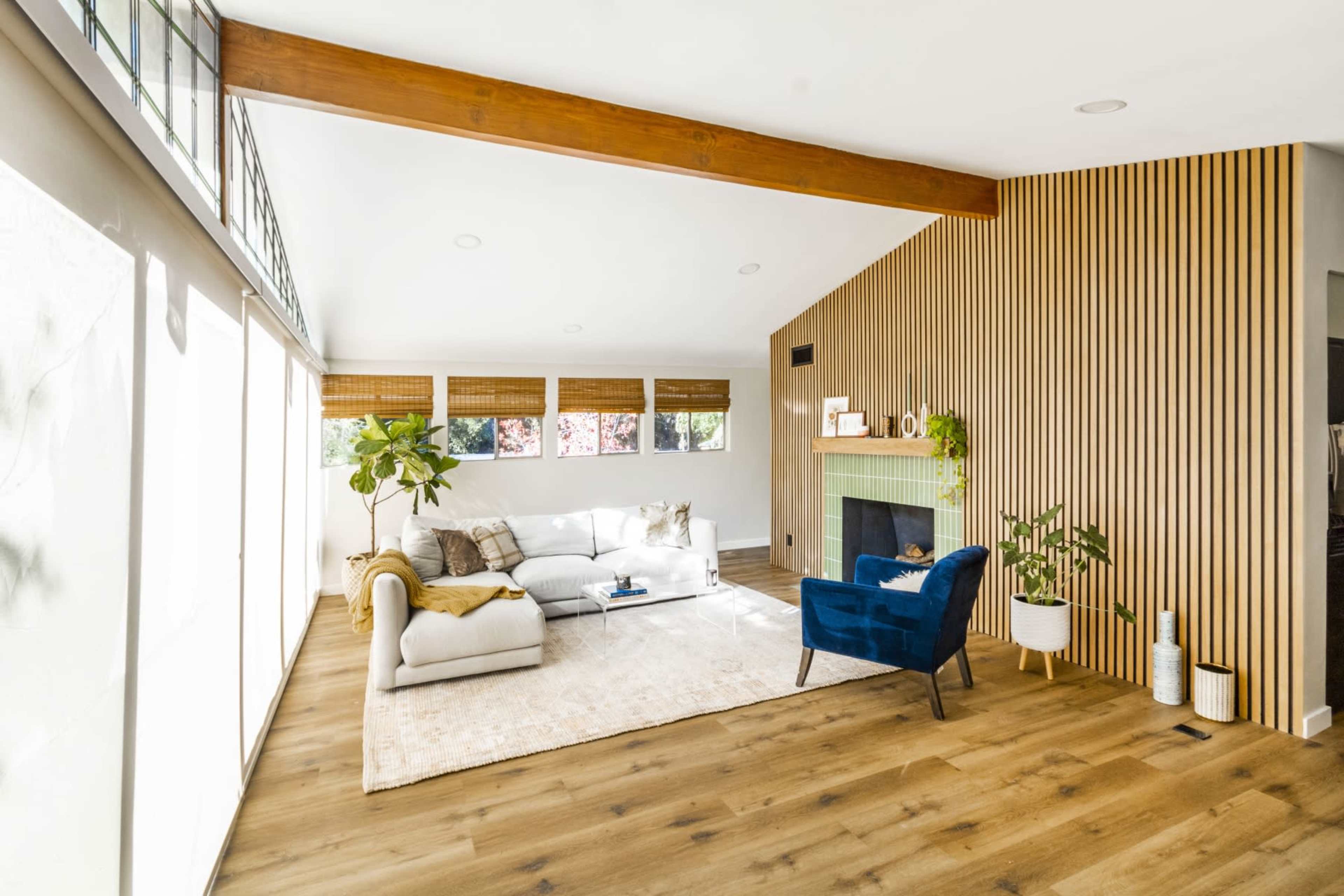 A modern living room features a light-colored sectional sofa, a blue armchair, a plant, and a fireplace, with large windows allowing natural light to enter.