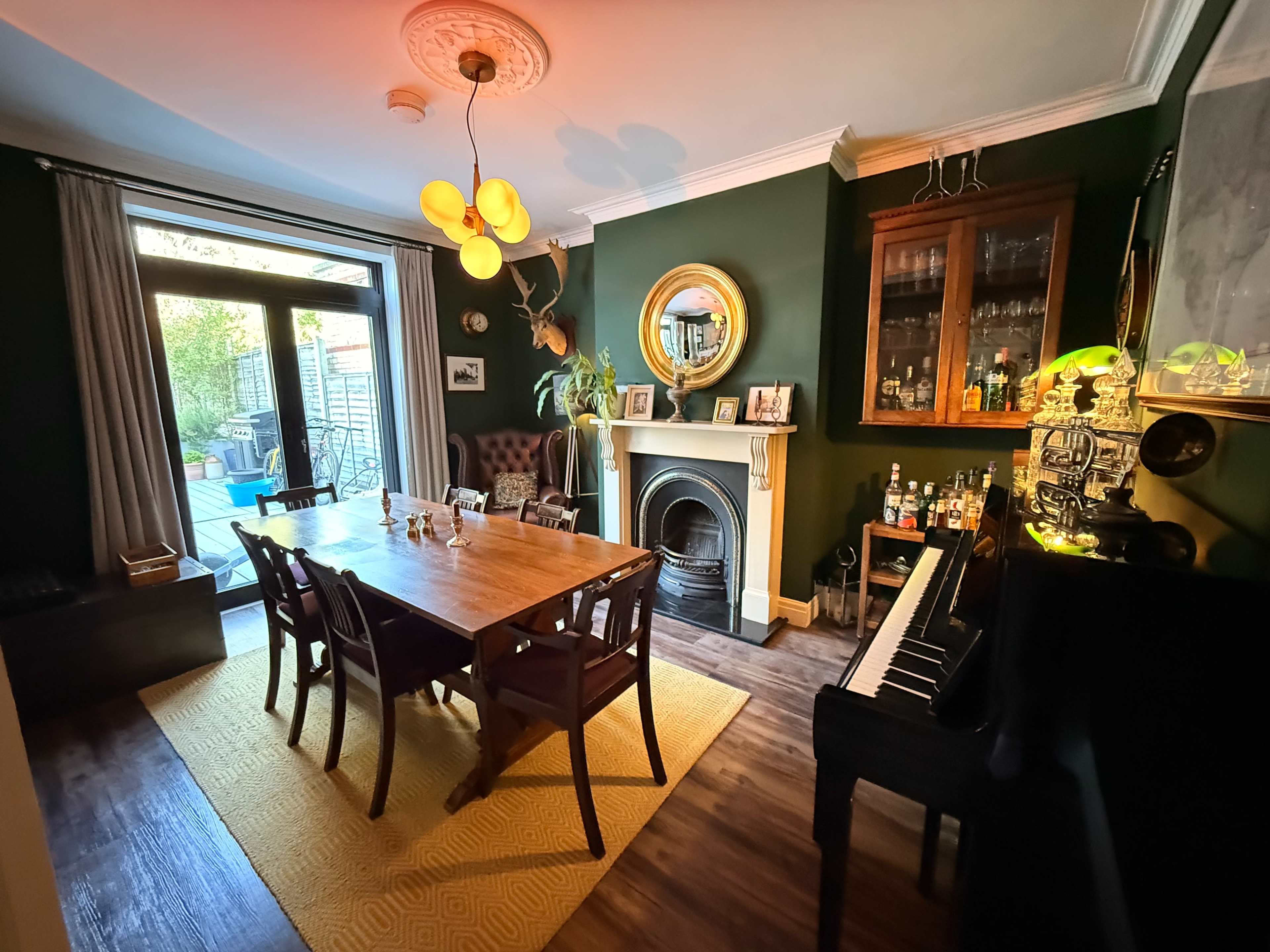 The image shows a dining room featuring a wooden table with chairs, a fireplace, and a piano, illuminated by pendant lights and natural light from a glass door.