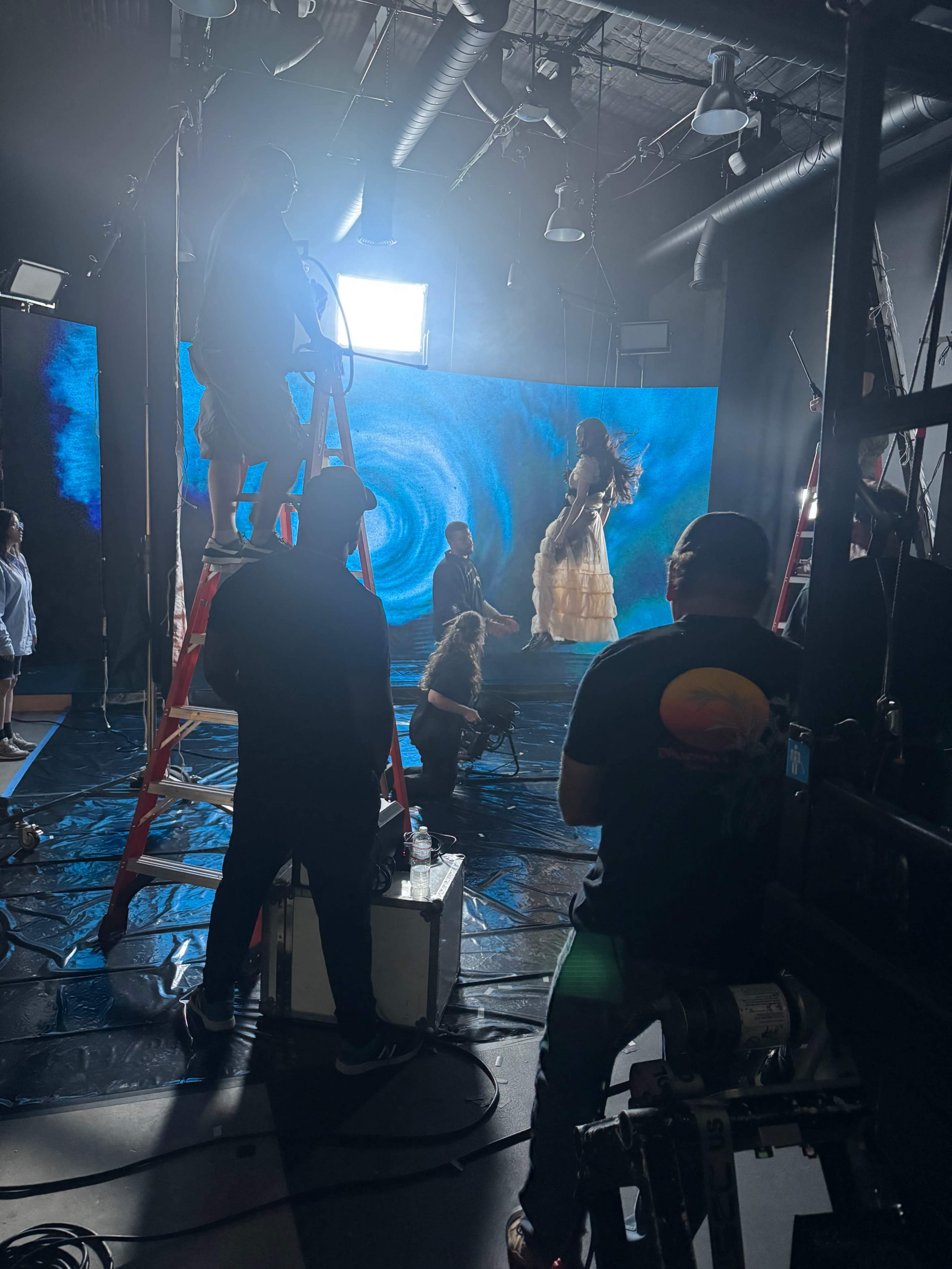 A production crew works on a set with large screens displaying a swirling blue backdrop while a performer in a dress is positioned for a shoot.